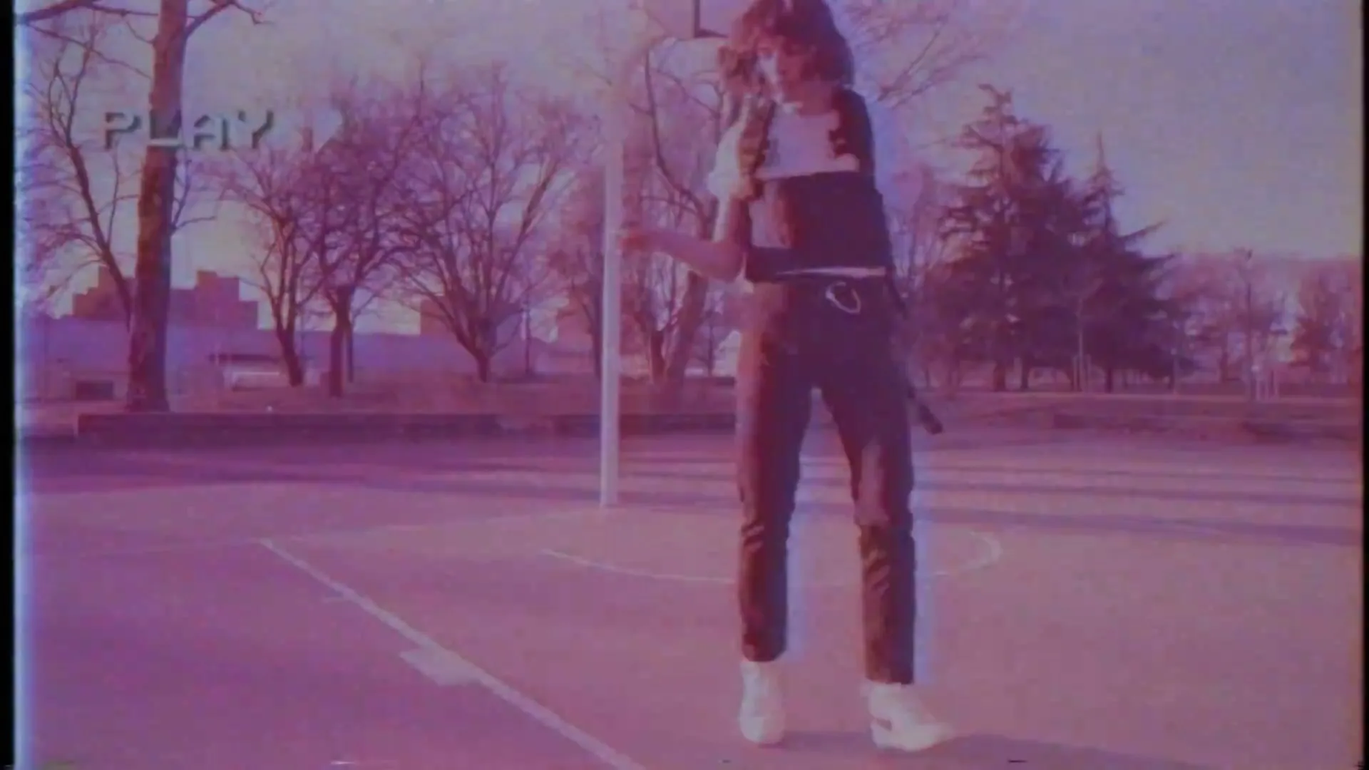 A young person with long hair and a vest dances on an outdoor basketball court, with trees in the background, filmed with a vintage camcorder effect.