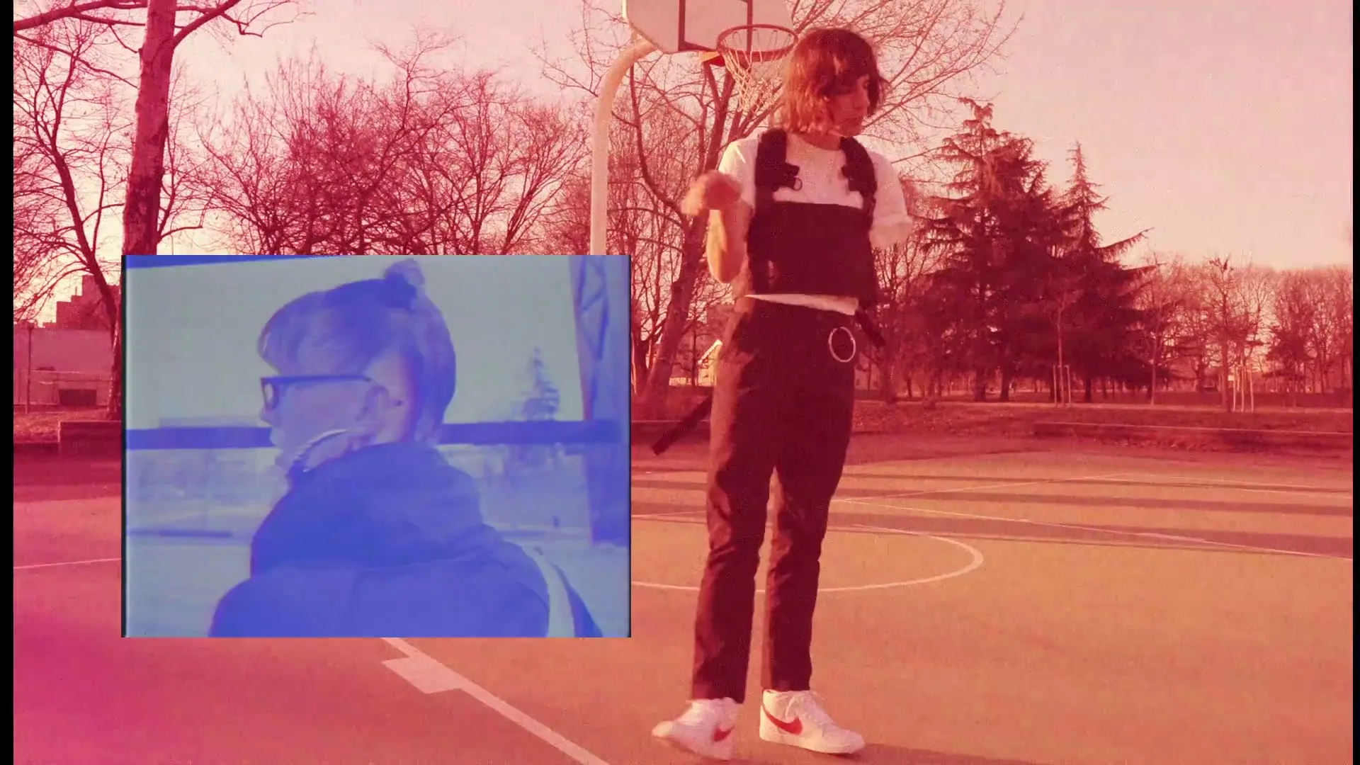 A person with long hair and a harness over a white t-shirt dances on a basketball court, with a blue-tinted video inset of another person.