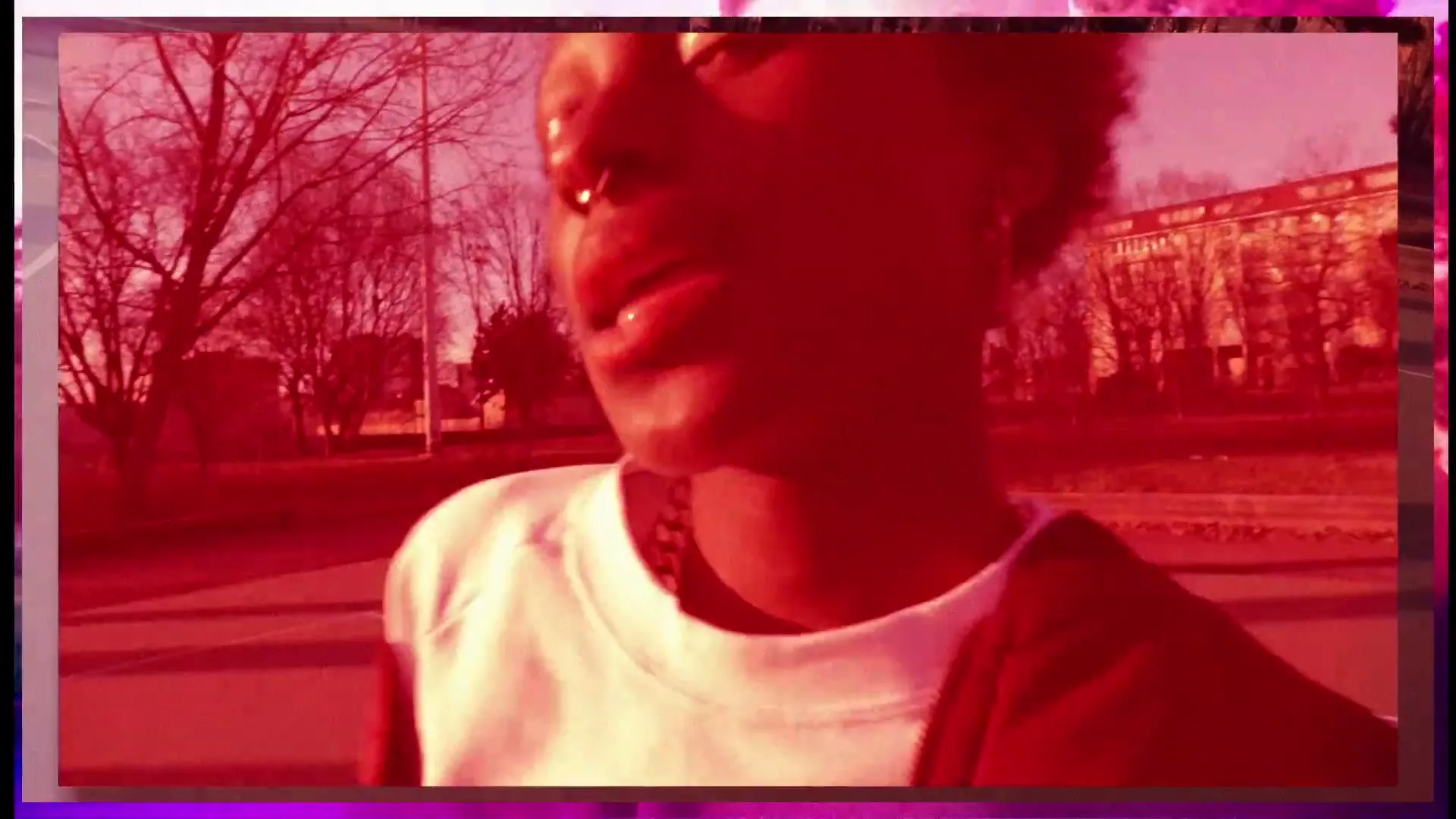 A young Black person with a nose ring and short curly hair looks at the camera, then away, on an outdoor basketball court.