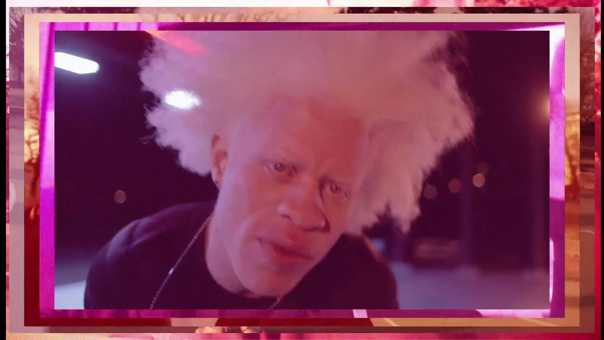 A man with white afro hair and light skin looks directly at the camera with a serious expression at night.