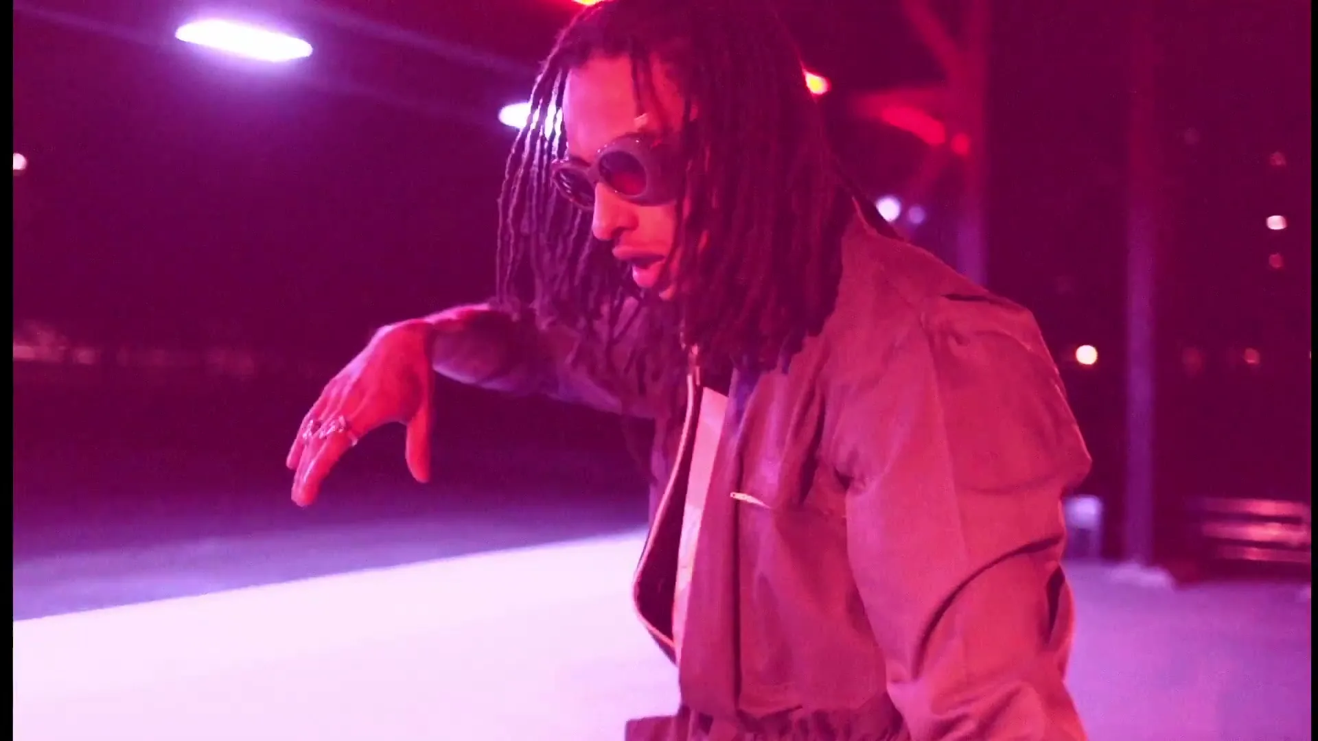 A young man with dreadlocks and sunglasses dances under neon lights in a dark, urban setting, illuminated by pink and purple hues.