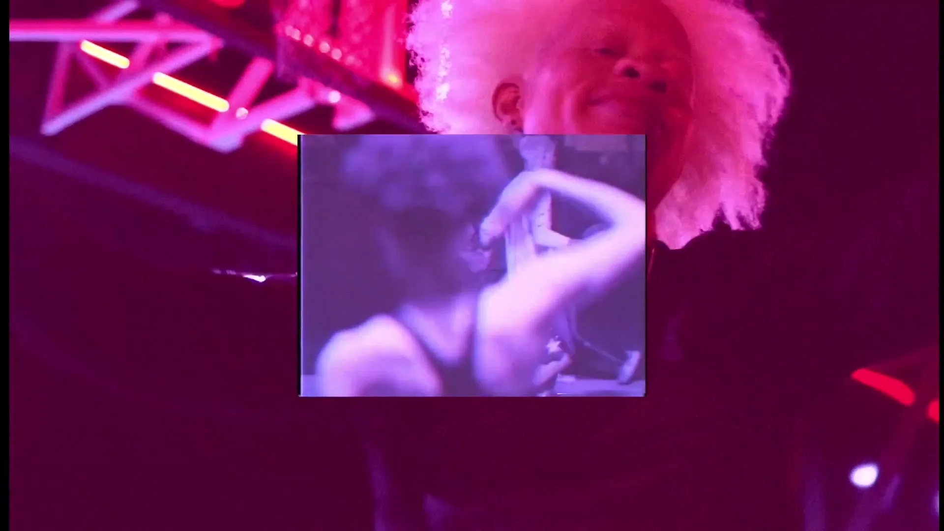 A person with white hair and a black shirt smiles, with a small video playing in the center of the frame, showing a person dancing.