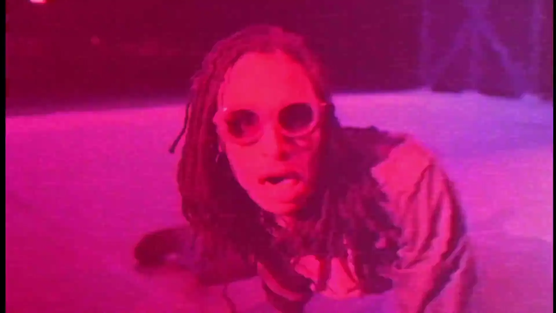 A person with dreadlocks and sunglasses crawls on the ground, looking up at the camera with an open mouth, in a pink and purple lit space.