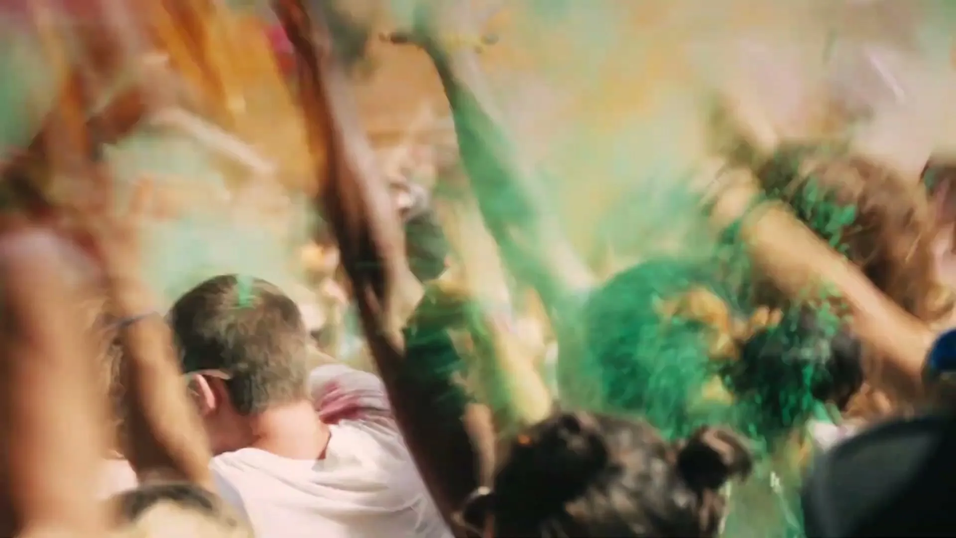 A crowd of young people at a festival throw colorful powder into the air, creating a vibrant, hazy atmosphere under the bright sun.