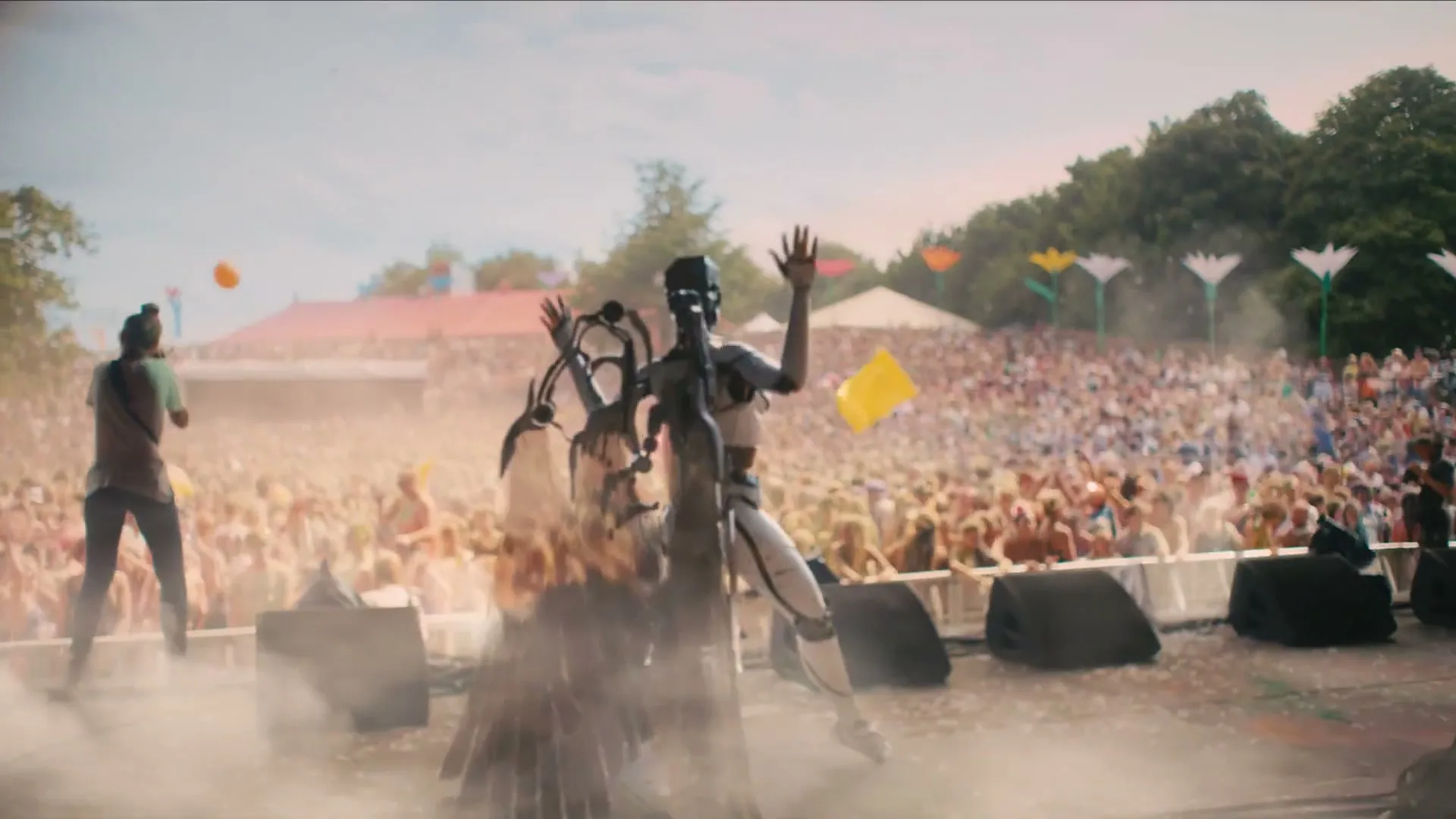 A robot with a flowing cape and a human with a ponytail dance on a stage, surrounded by a large crowd at an outdoor music festival.