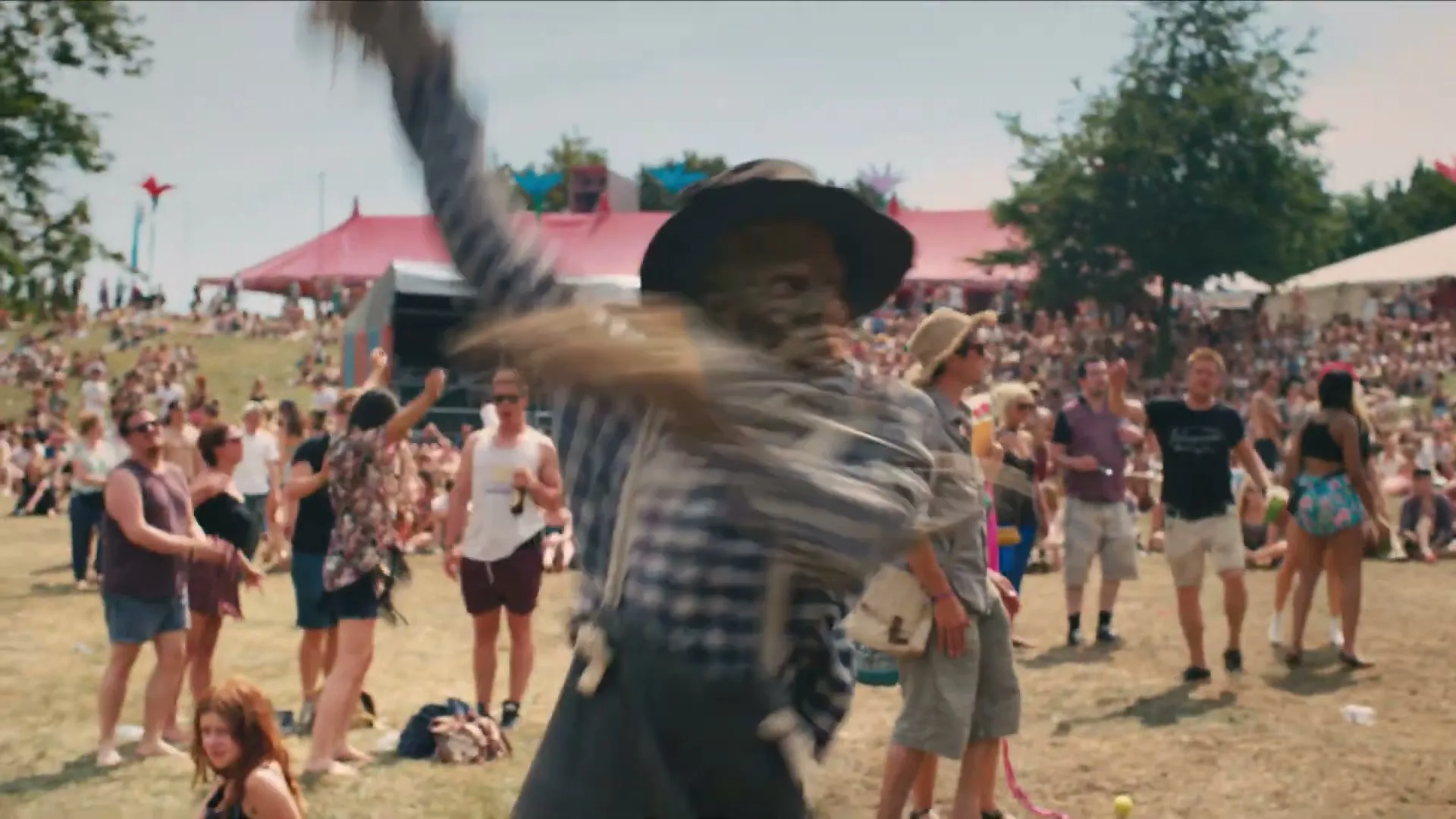 A scarecrow-like figure with a hat and plaid shirt dances energetically in a crowded outdoor music festival during the day.
