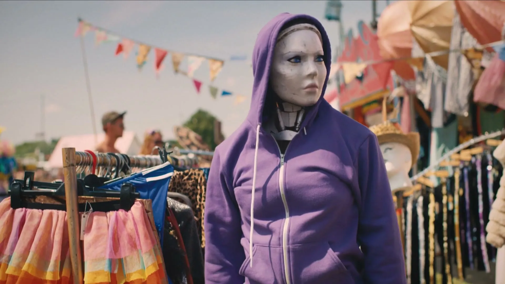 A robot with a human-like face and blue eyes, wearing a purple hoodie, looks around a bustling outdoor market filled with clothes and colorful bunting on a sunny day.