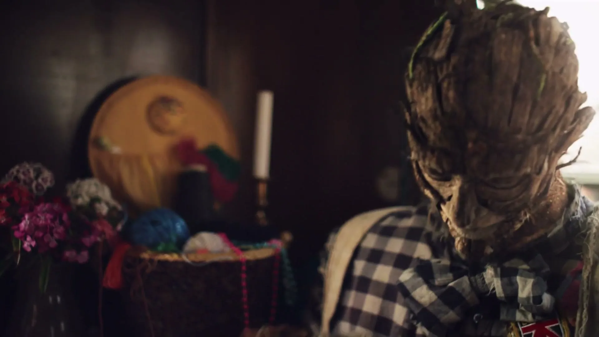 A scarecrow with a tree-like face and a plaid shirt looks around a dimly lit room with colorful decorations.