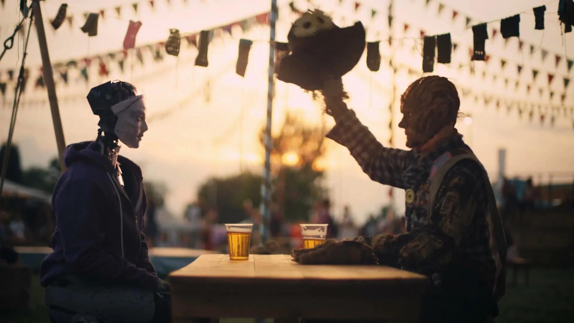 A robot and a scarecrow sit at a wooden table with two beers, as the scarecrow raises its hat in a festive outdoor setting at sunset.