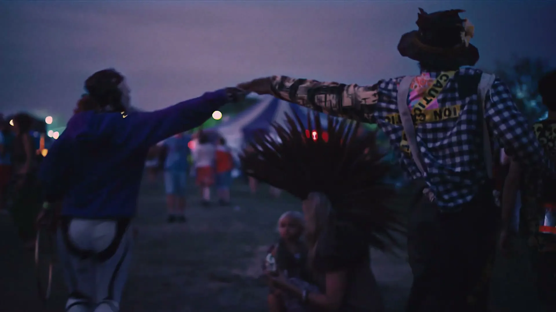 Two people in costumes, one dressed as a robot and the other as a scarecrow, dance together at a festival at dusk.