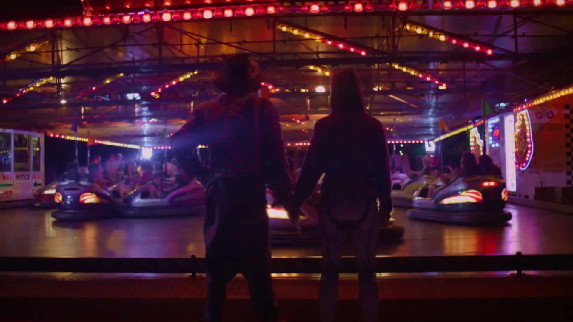 A scarecrow and a robot, holding hands, stand with their backs to the camera, watching bumper cars at a brightly lit fairground at night.