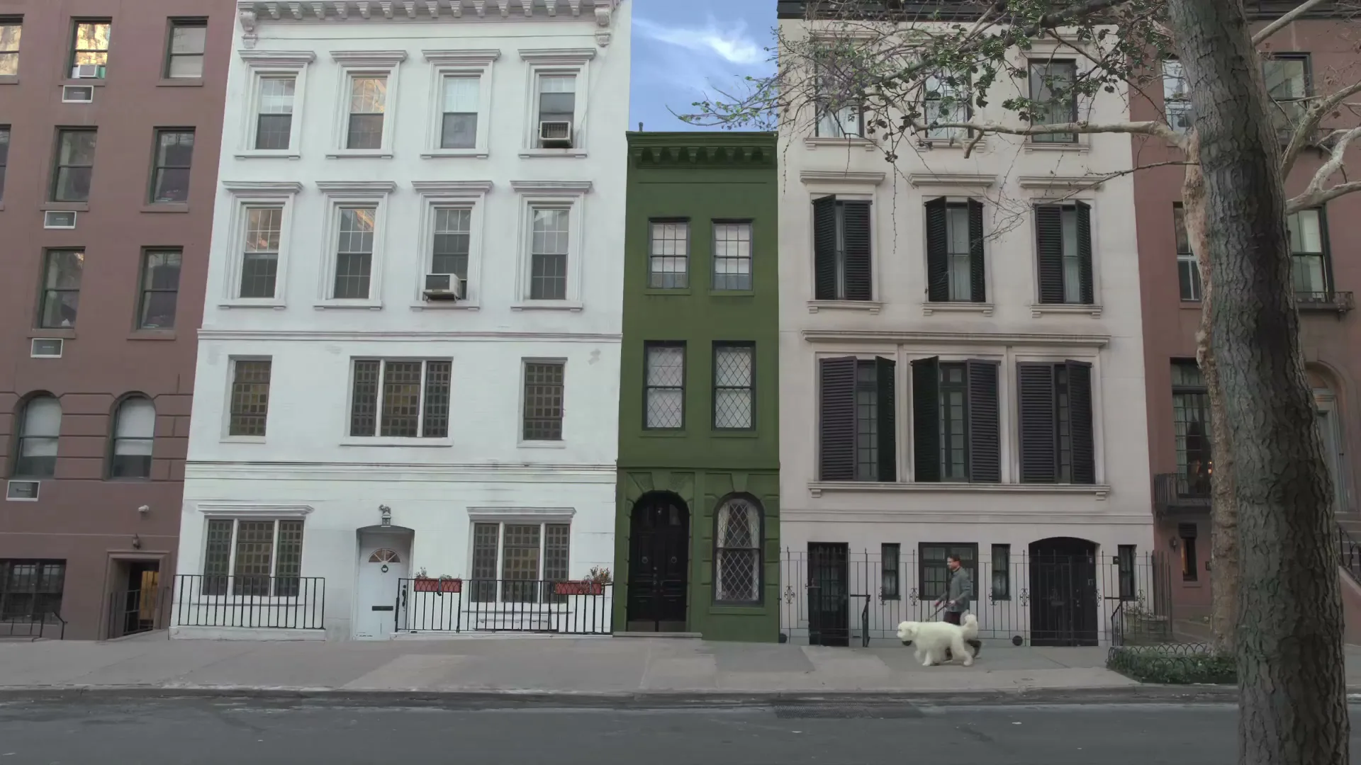 A man walks a white dog past a row of townhouses, one of which is painted green, on a city street during the day.