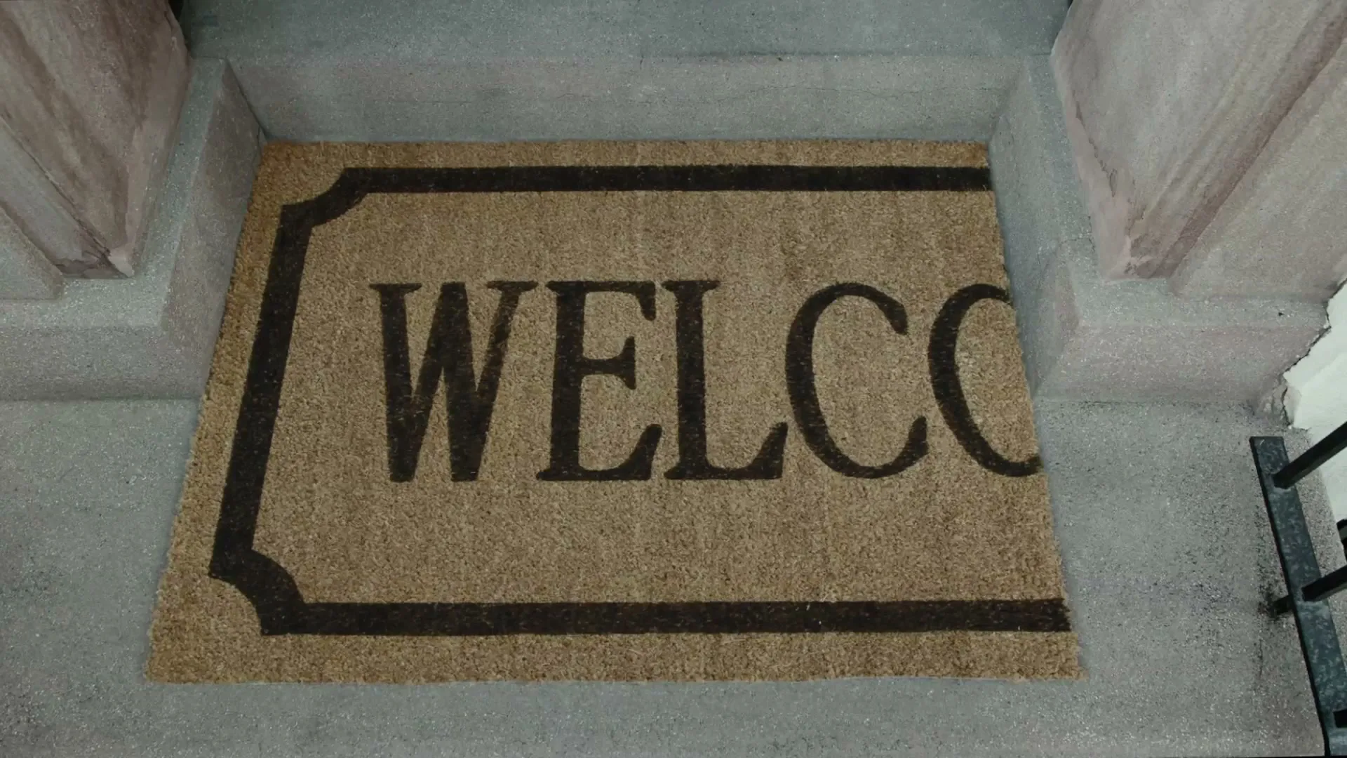A brown doormat with the word "WELCO" printed in black is seen on a concrete doorstep, with a black railing visible on the right.