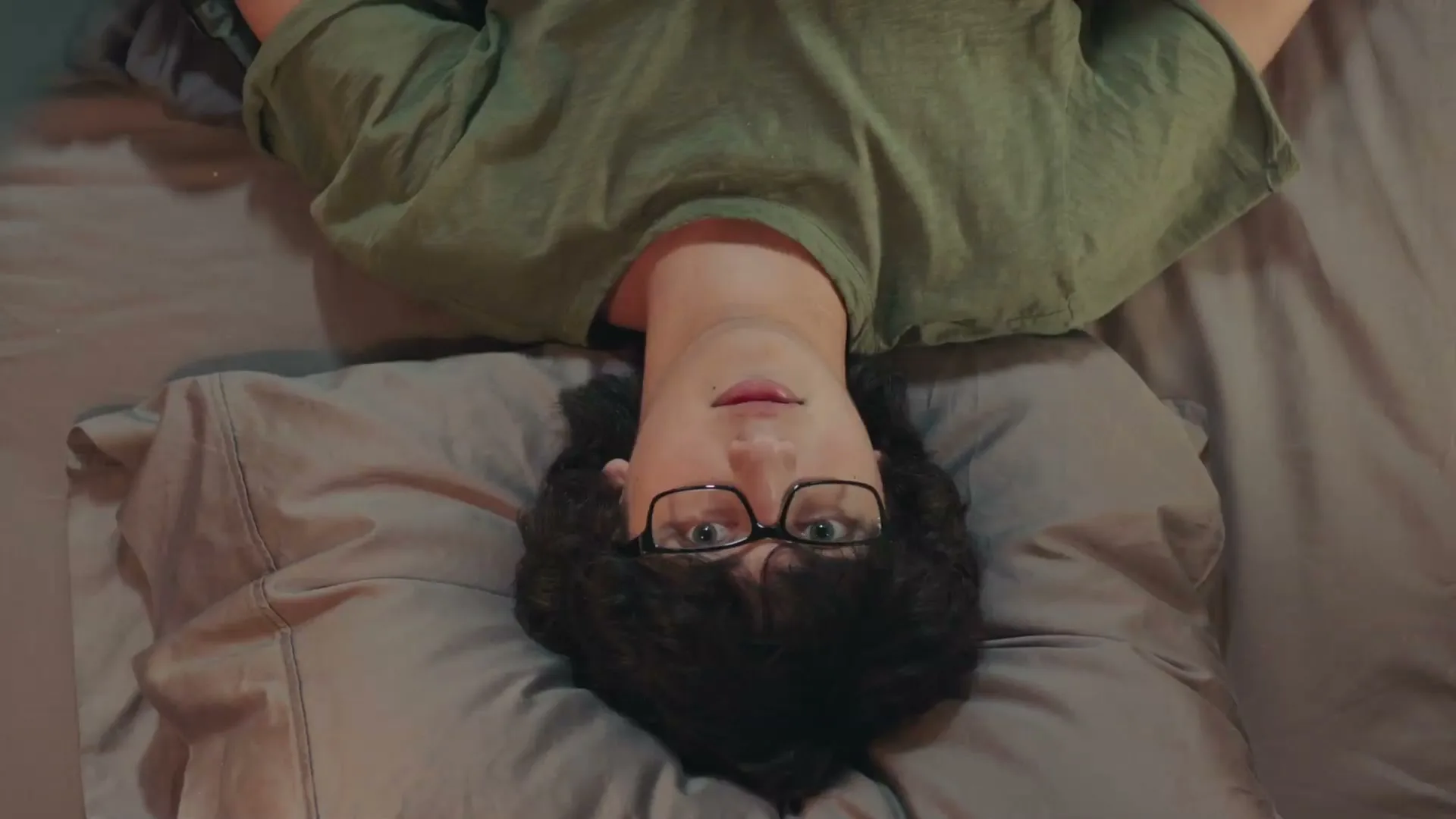A young man with dark curly hair and glasses lies upside down on a bed, looking directly at the camera with a neutral expression.
