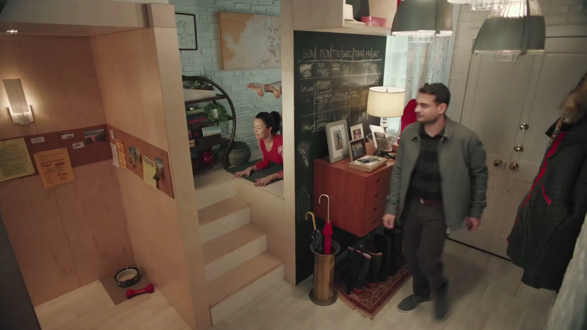 A man walks into a small apartment, while a woman practices yoga in a lofted area and a dog runs around.