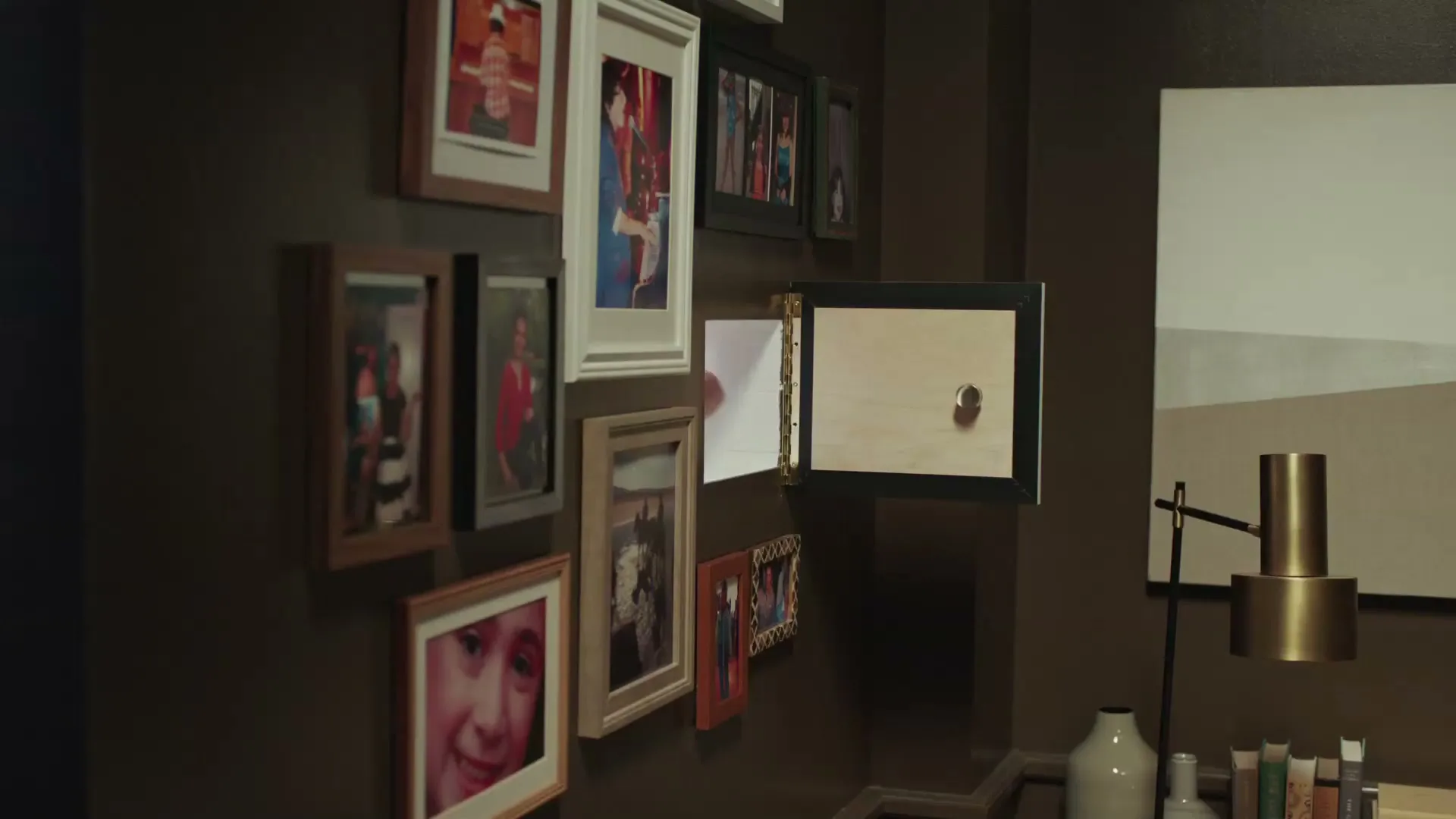 A hand reaches through a picture frame on a wall filled with family photos, as if passing through a portal, in a home interior.