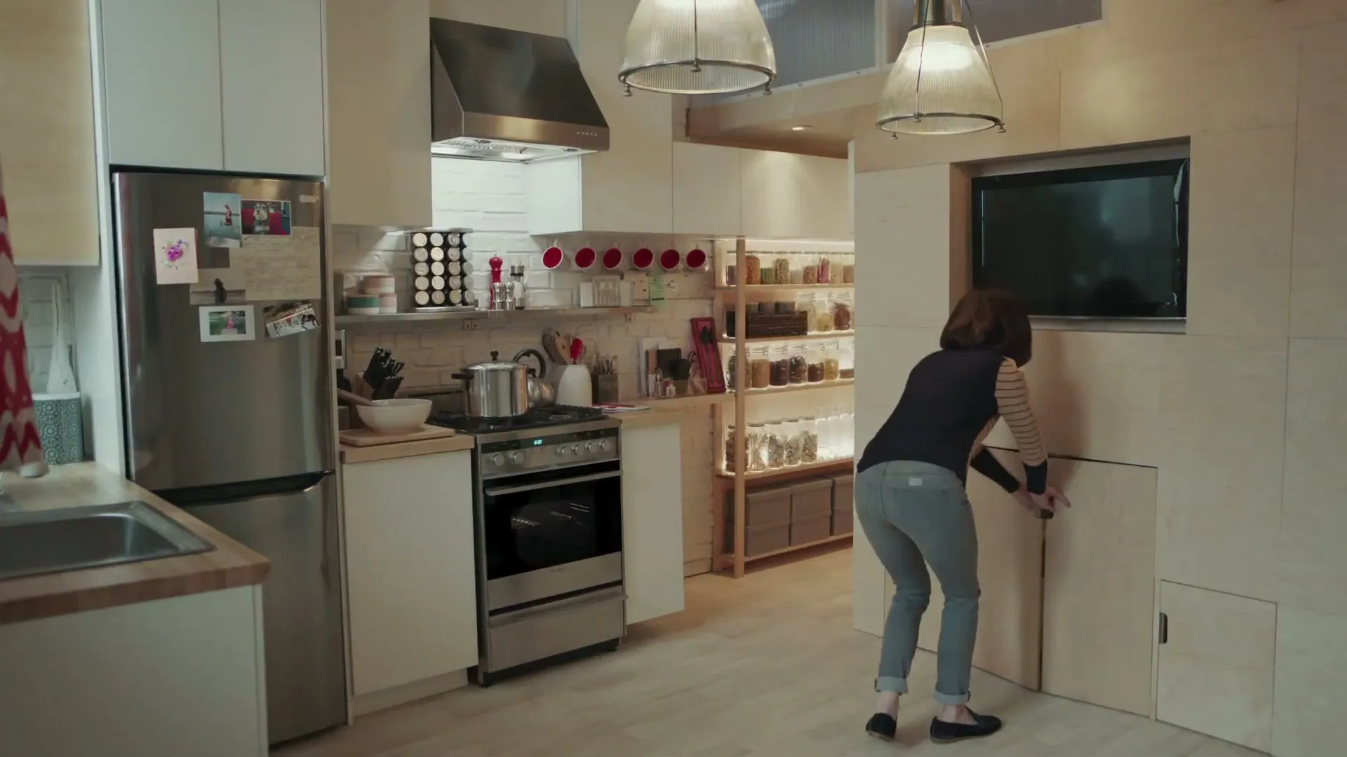 A woman in a striped shirt and jeans walks into a modern kitchen, opens a hidden compartment, and pulls out a folding chair.