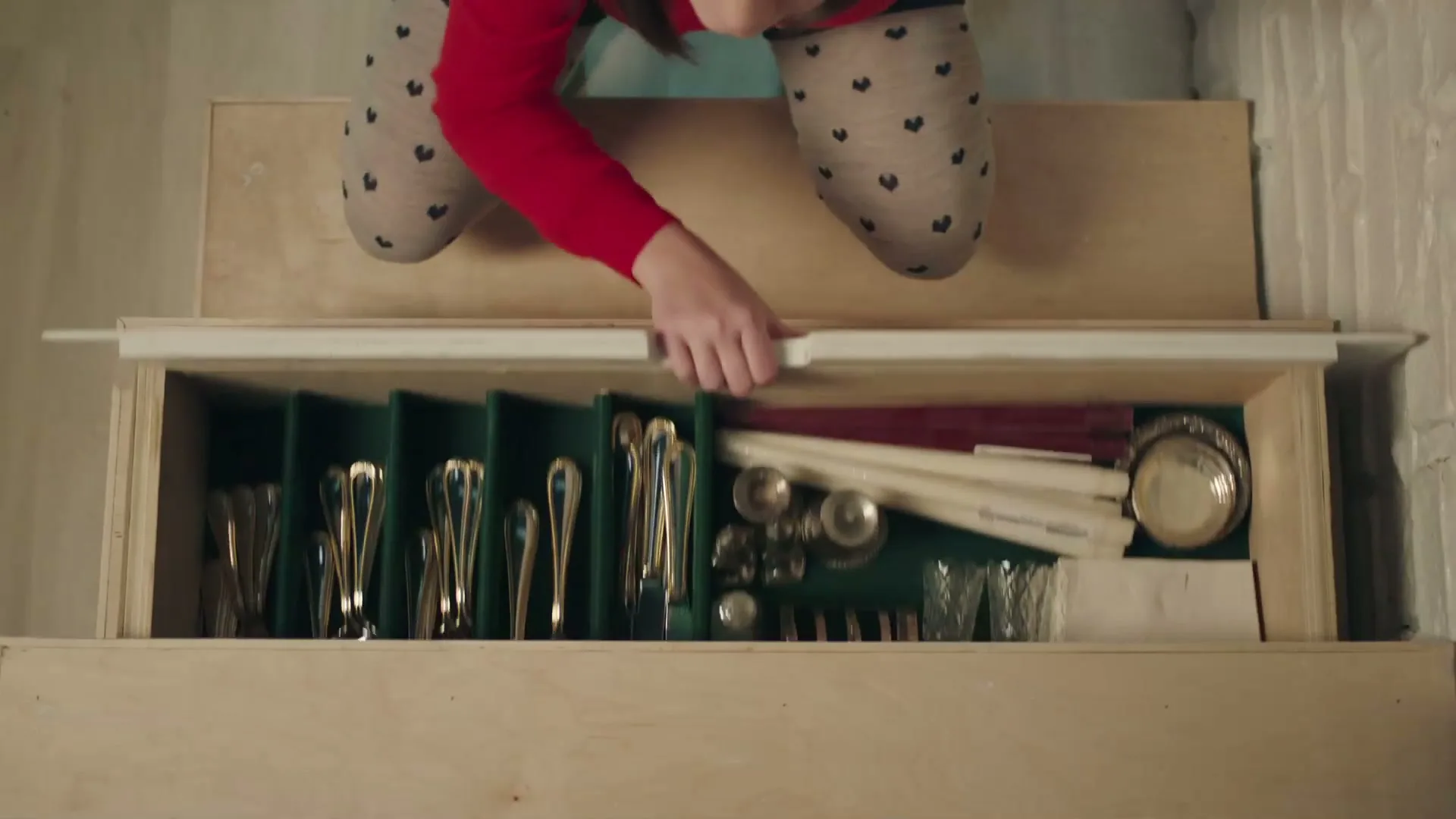 A young girl in a red shirt and patterned leggings opens a hidden drawer in a wooden staircase, revealing silverware and other items.