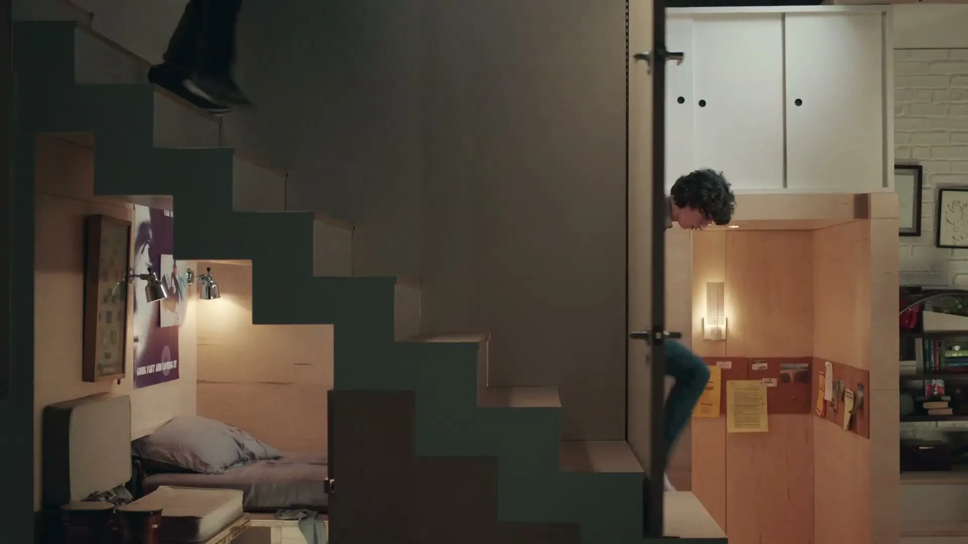 A man and a dog walk down a set of stairs in a small, compact apartment, while a child peeks out from a small room below the stairs.