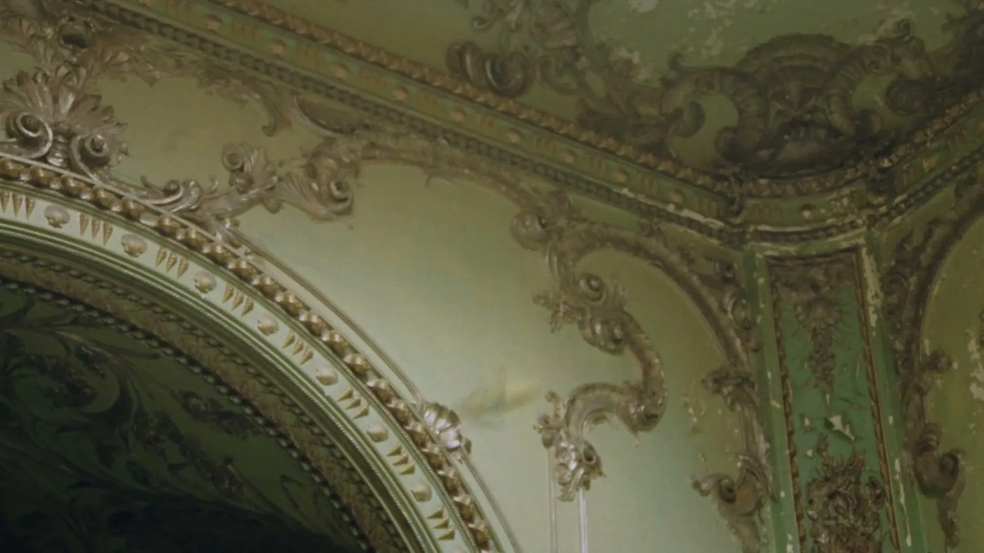 A detailed shot of an ornate, decaying wall and ceiling with intricate gold and green patterns in an old, abandoned building.
