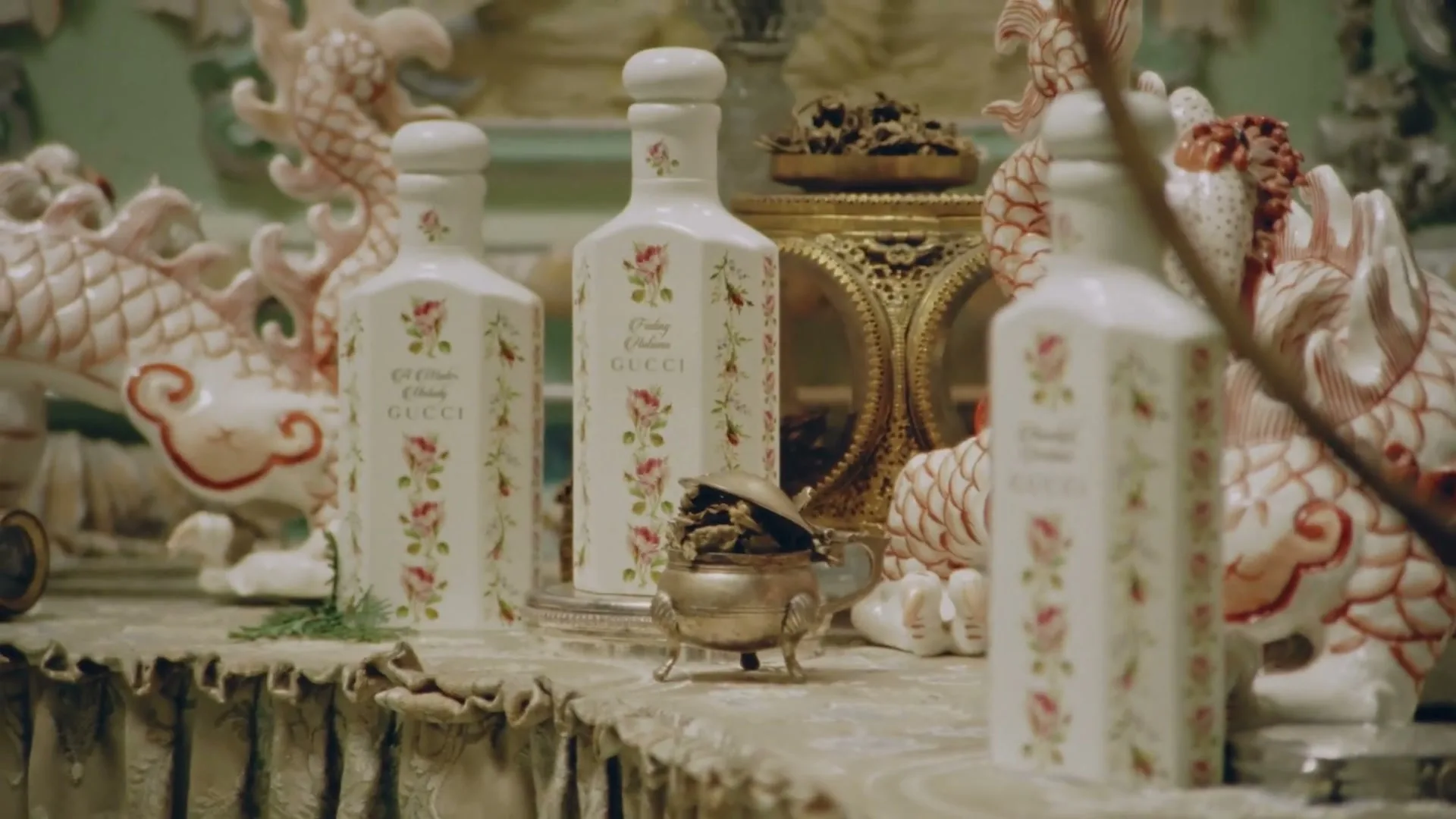 Three white, floral-patterned perfume bottles stand on a decorative table, surrounded by ornate porcelain dragons and a small silver incense burner.