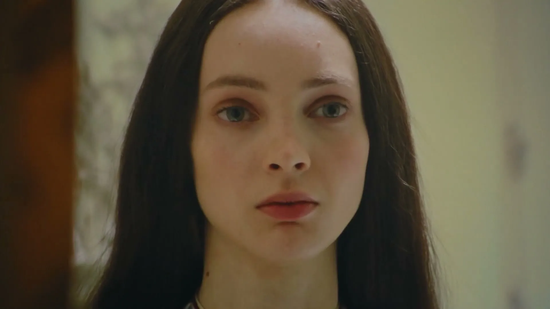 A young woman with long dark hair and a white dress with a red and blue pattern looks directly at the camera, then slowly closes her eyes in an interior setting.