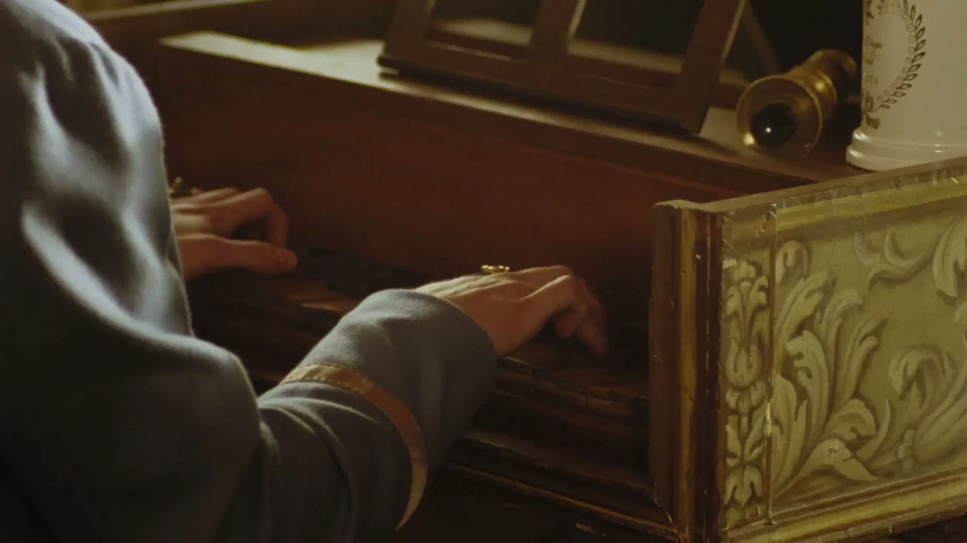 A person in a grey uniform with gold trim plays a harpsichord, their hands moving across the keys in a dimly lit room.