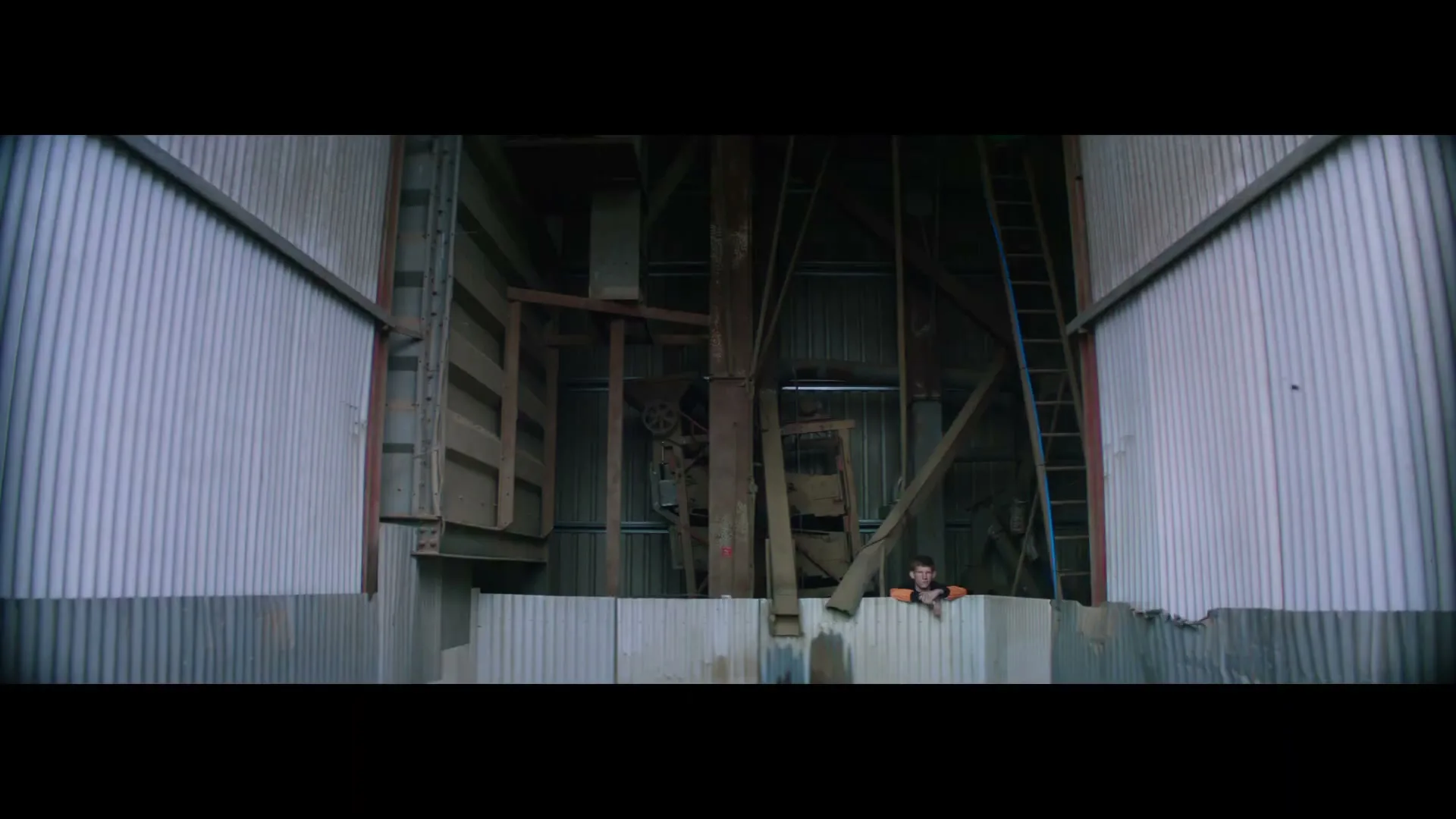 A young man in an orange shirt looks over a corrugated metal wall inside a large, industrial building with wooden beams and machinery.
