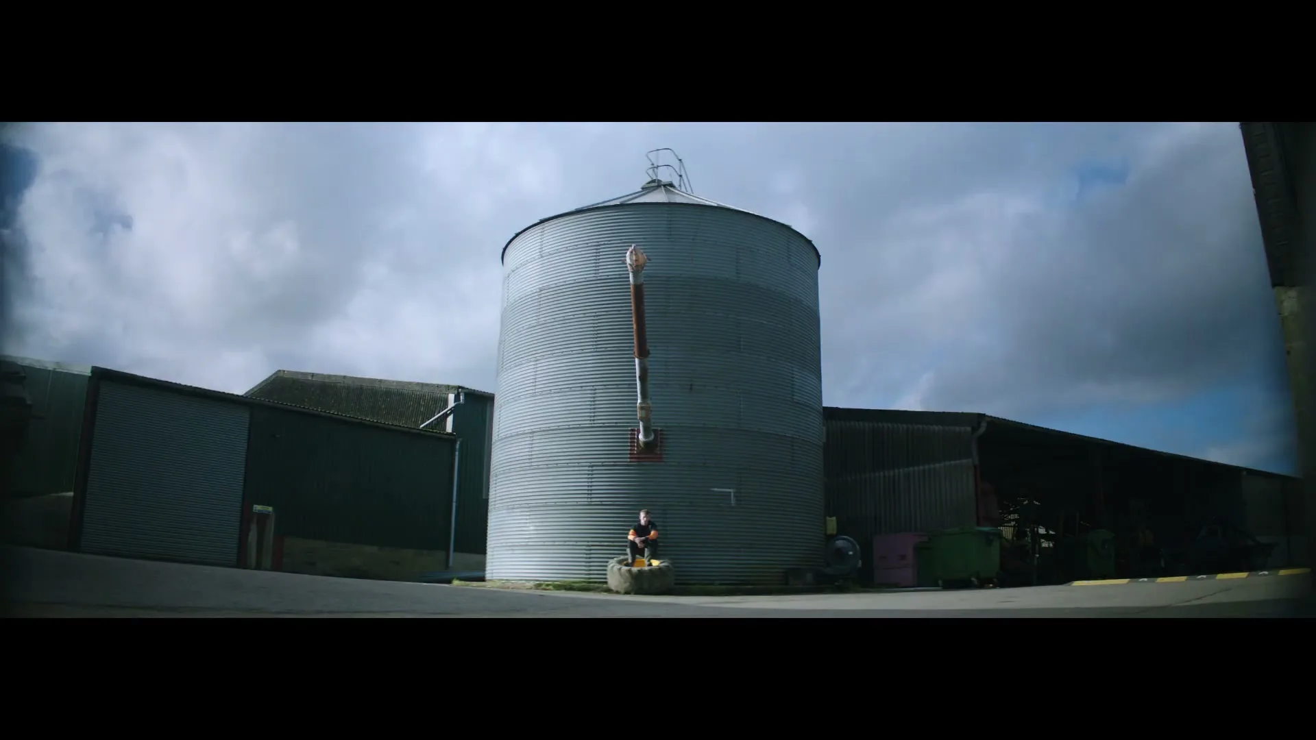 A young man in a black t-shirt and shorts sits on a tire in front of a large metal silo on a farm under a cloudy sky.