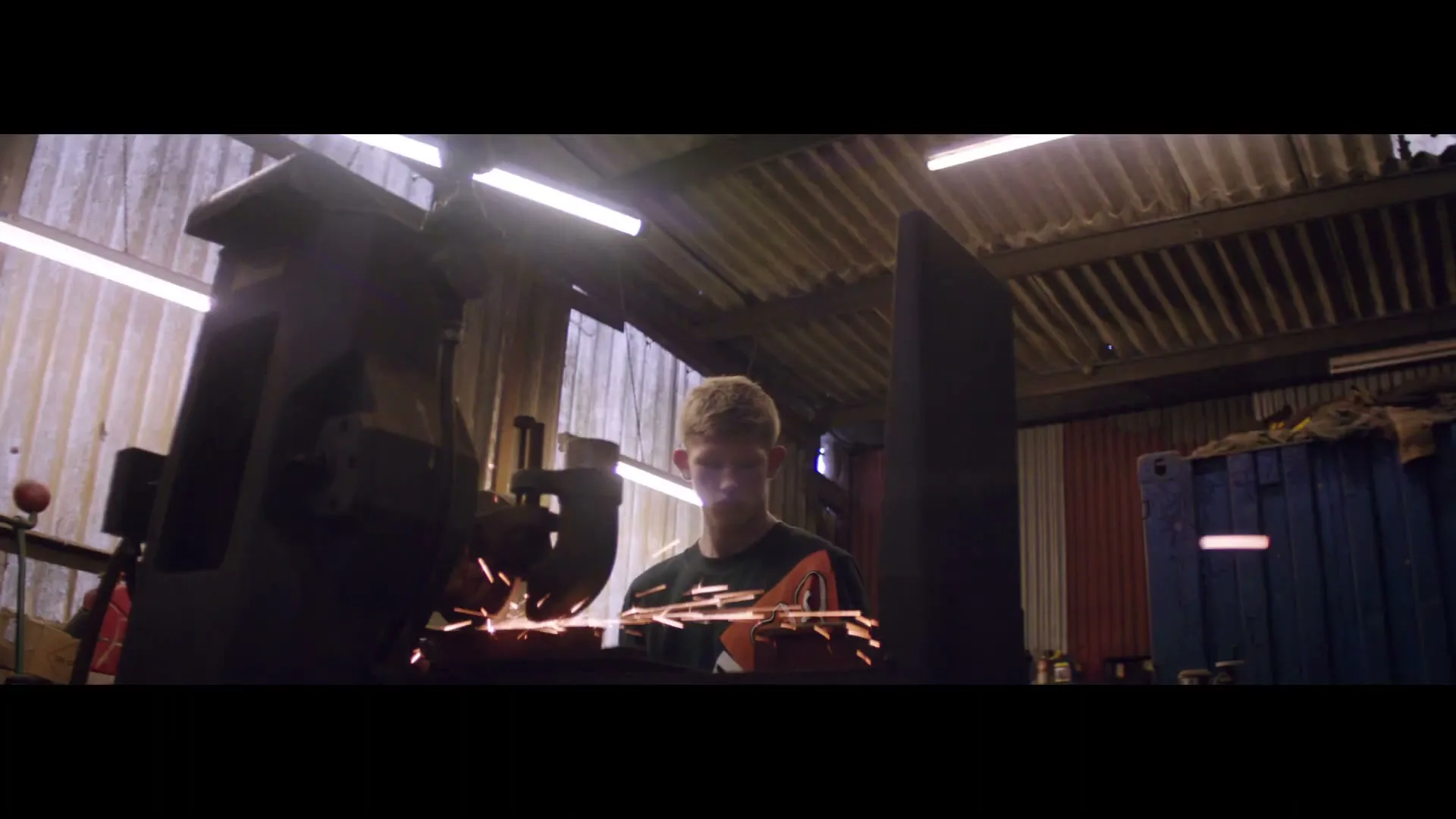 A young man with short blonde hair, wearing a dark green shirt, is focused on grinding metal, creating sparks in a dimly lit workshop.