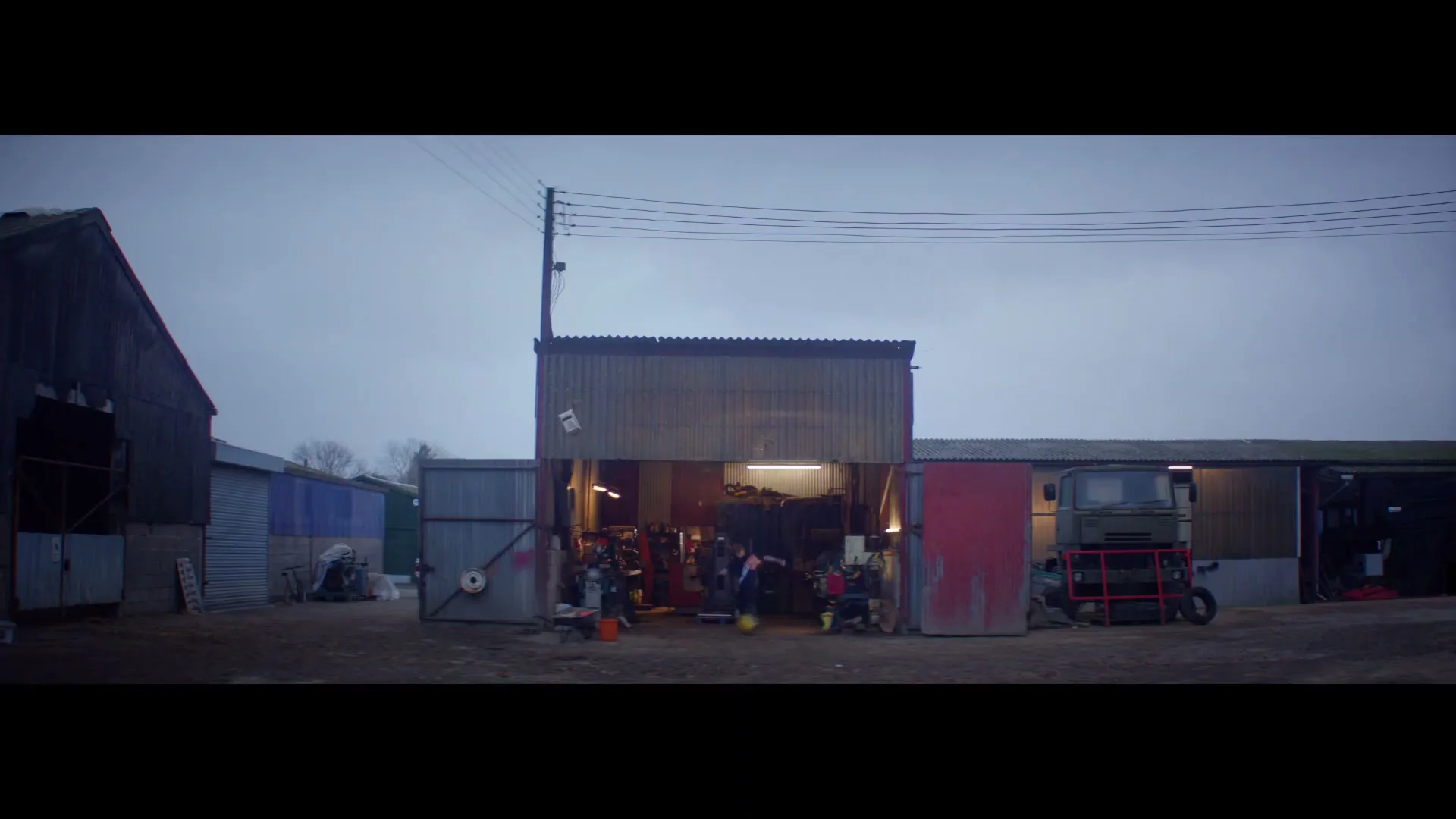 A wide shot shows a group of young men in casual clothes running out of a brightly lit garage on a cloudy day in a rural setting.