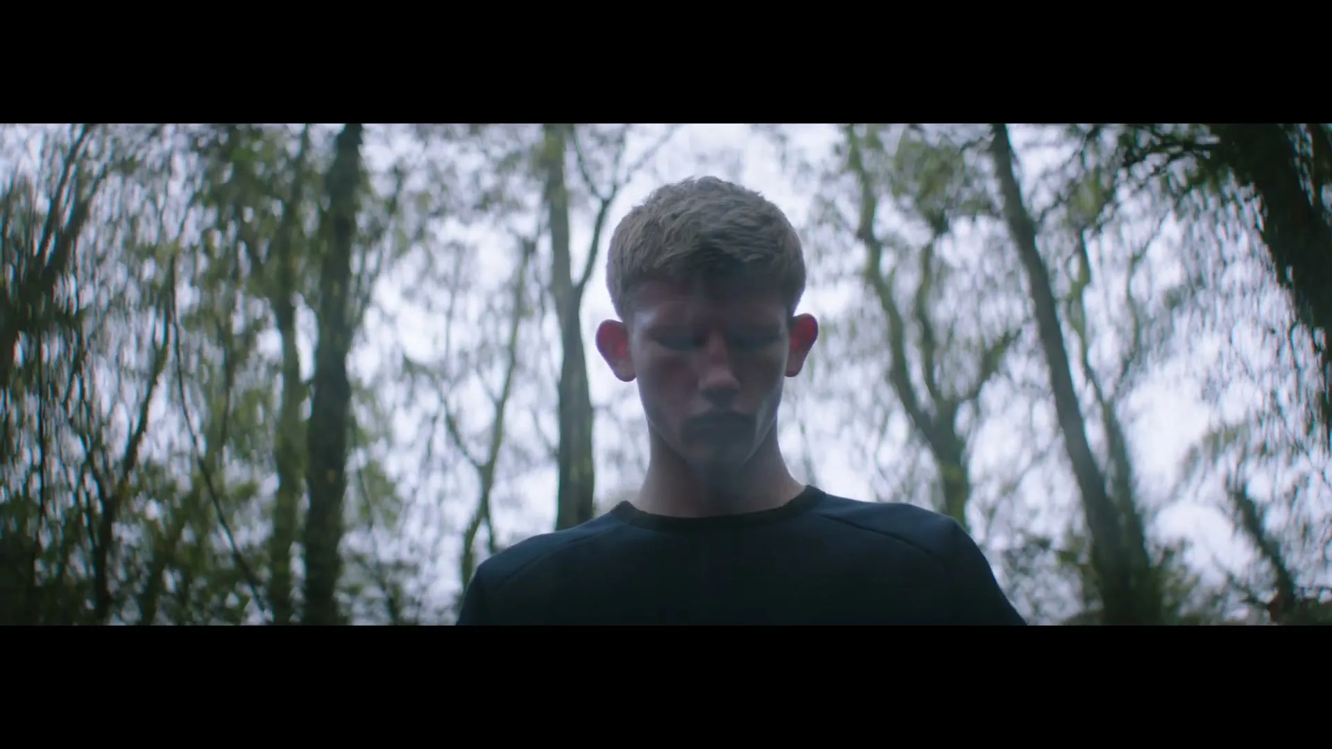 A young man with short hair and a dark shirt looks up in a forest with tall, bare trees against a bright sky.