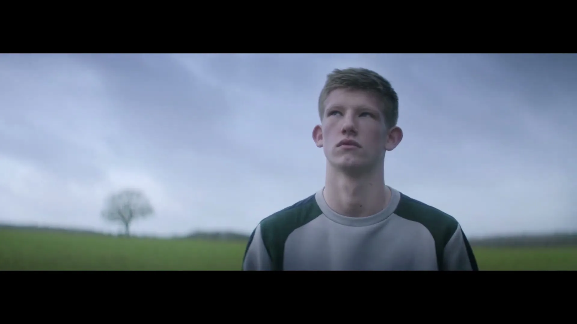A young man with short brown hair and a grey and green sweatshirt stands in a field with his eyes closed, then opens them and looks up.