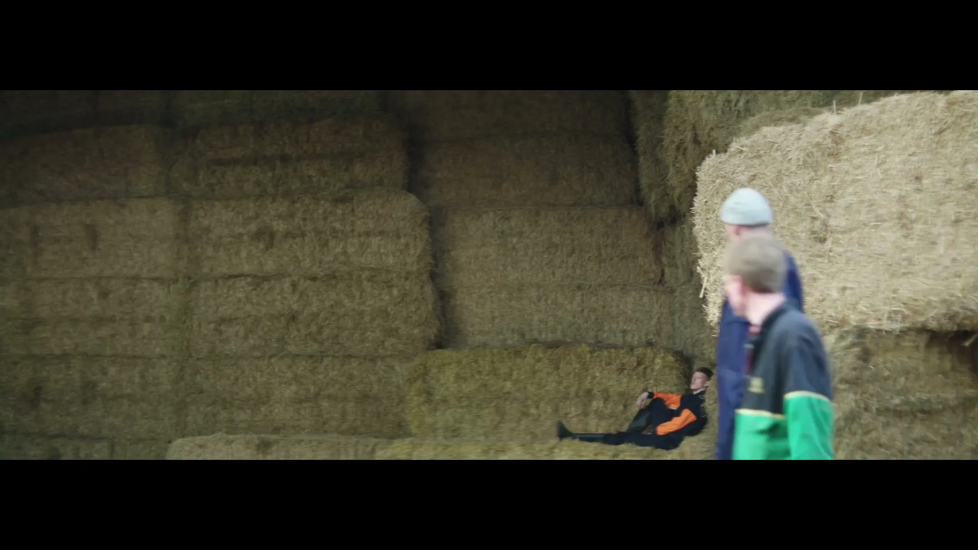 A young man in an orange and black tracksuit lies on a bale of hay as two older men walk past him in a barn.