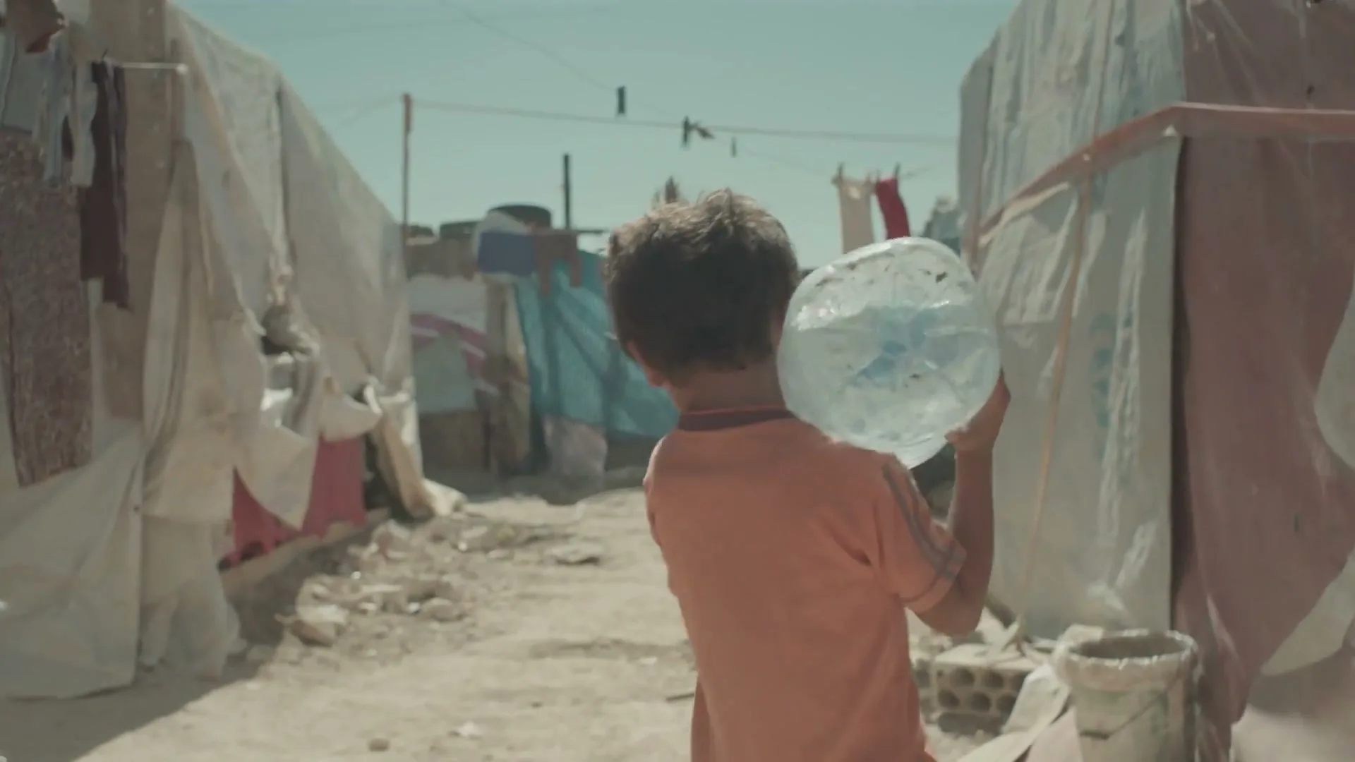 A young boy with curly hair, wearing an orange t-shirt, walks away from the camera carrying a large plastic water bottle through a dusty refugee camp.