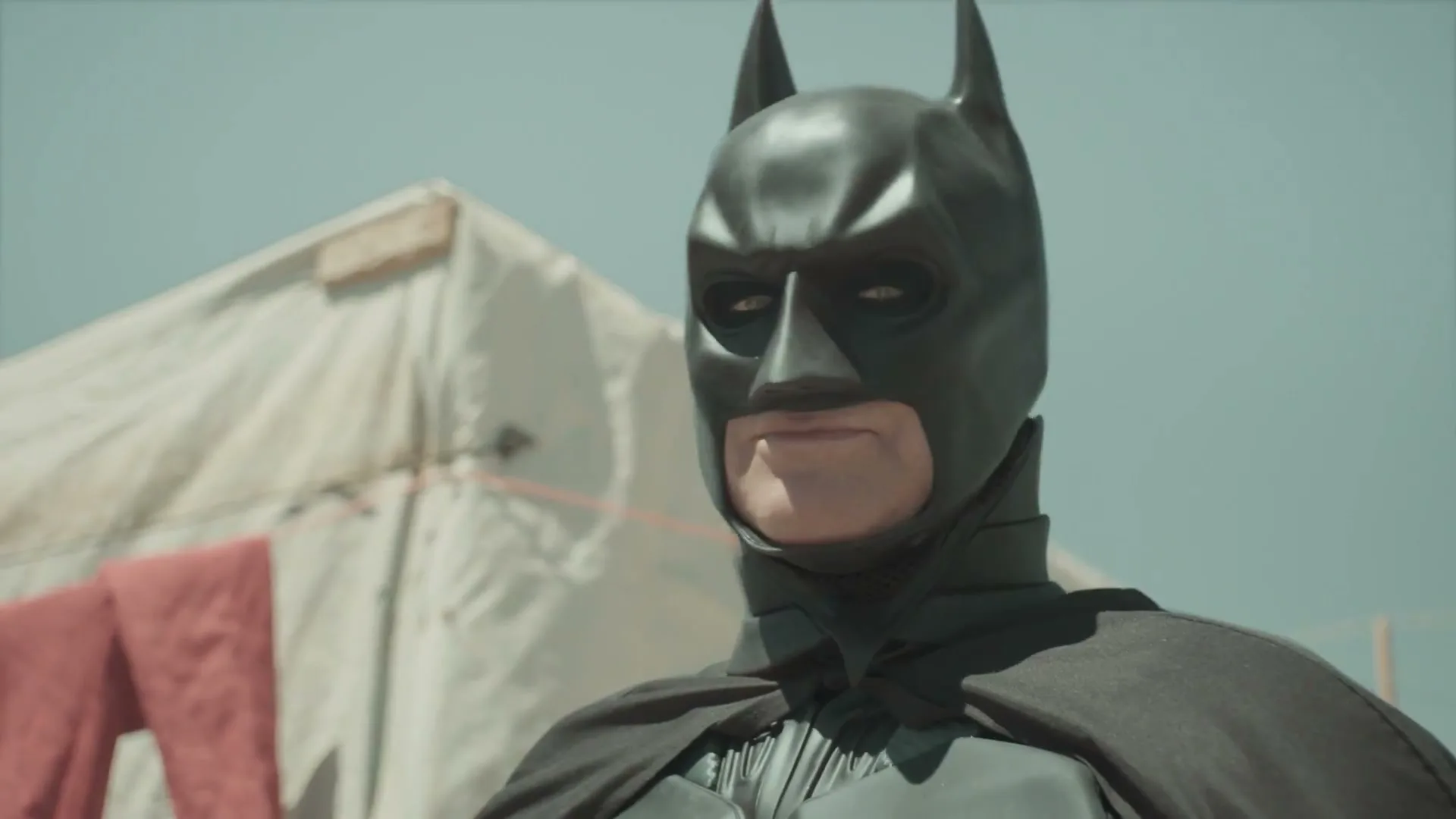 A person dressed as Batman stands in front of a white tent in a dusty, outdoor setting, looking off-camera with a serious expression.