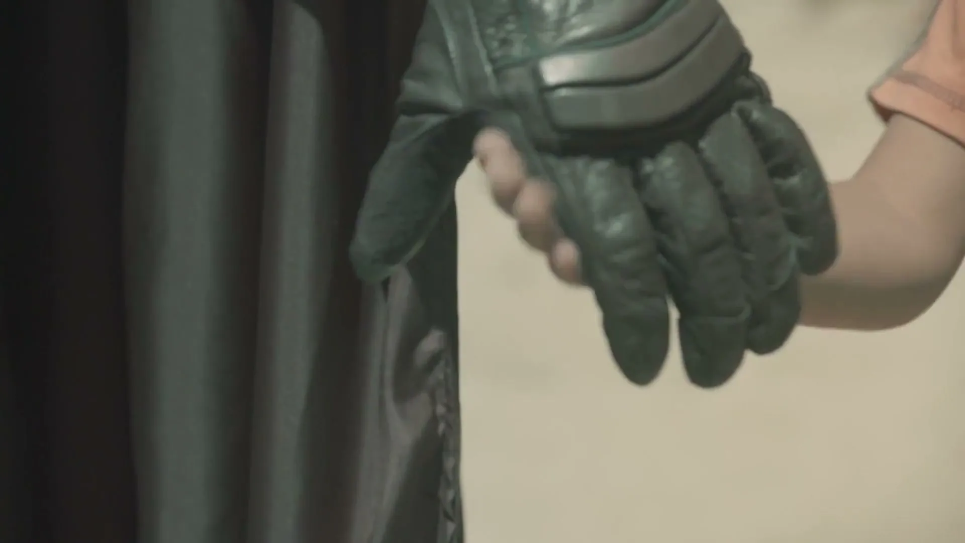 A child's hand reaches out and holds the gloved hand of an adult dressed in a dark costume, in an interior setting.