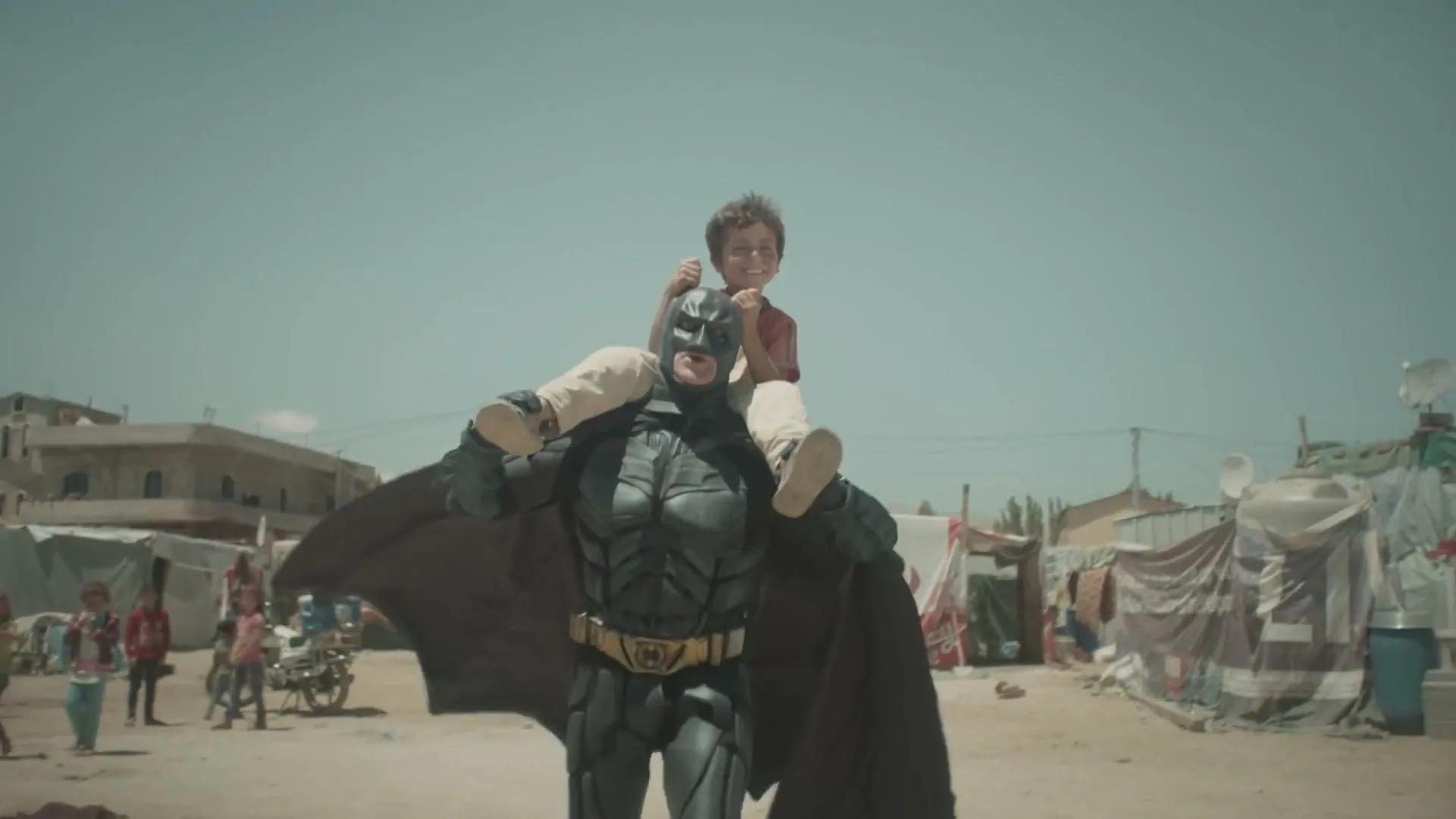 A man in a Batman costume carries a smiling child on his shoulders, walking through a dusty refugee camp under a bright sky.