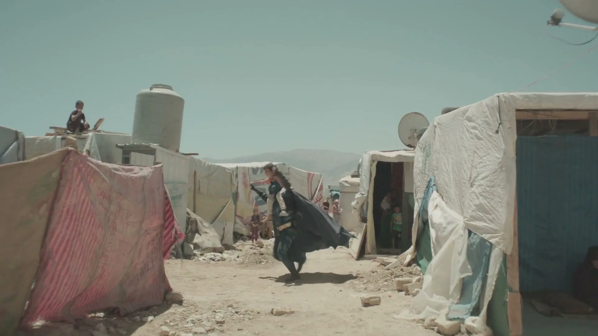 A man dressed as Batman carries a child on his shoulders, running through a dusty refugee camp under a bright sky.