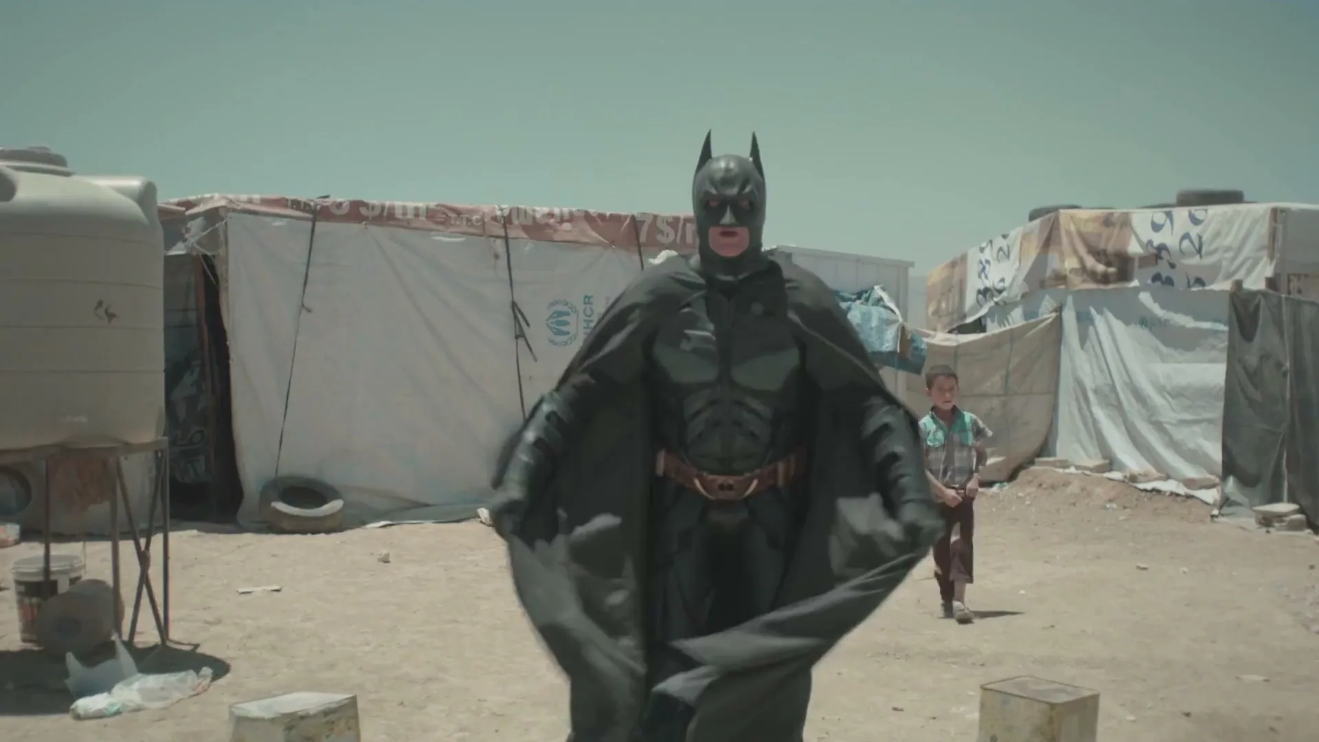 A man in a Batman costume stands in a refugee camp, spreading his cape as a child watches in the background.