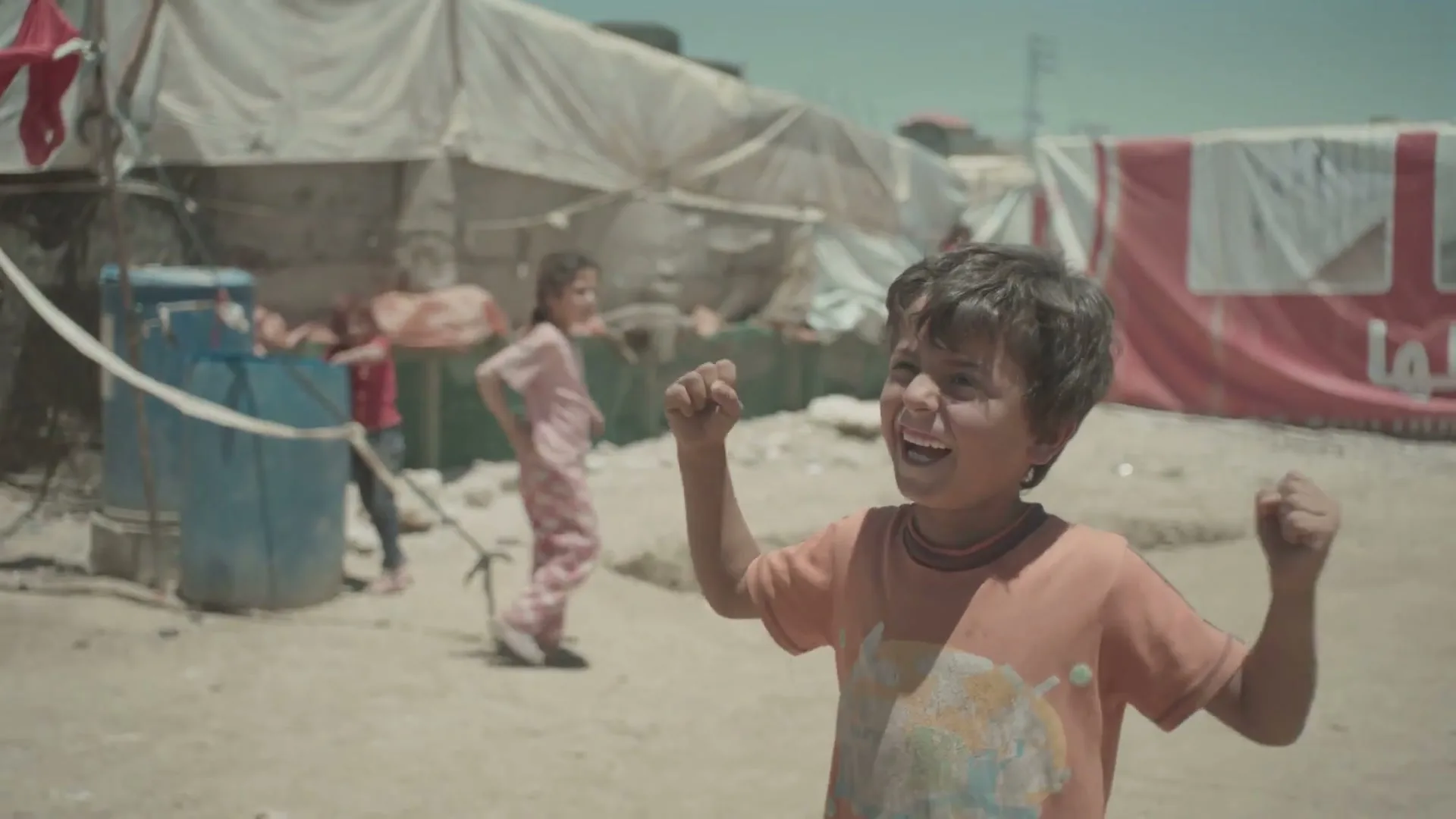 A young boy with dark hair and an orange t-shirt laughs joyfully, raising his fists in a refugee camp under a bright sky.
