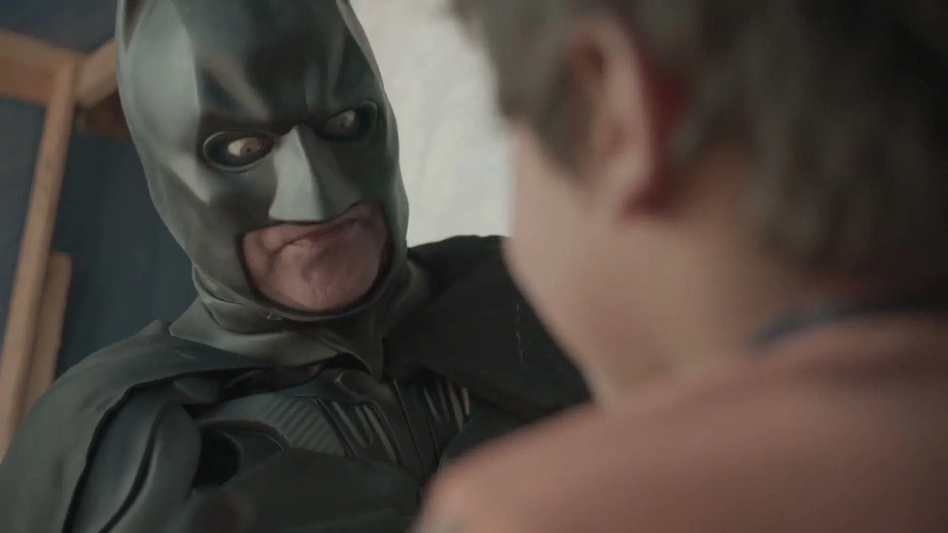 A man dressed as Batman looks down at a child from a low angle, with the child's back to the camera, in a dimly lit room.