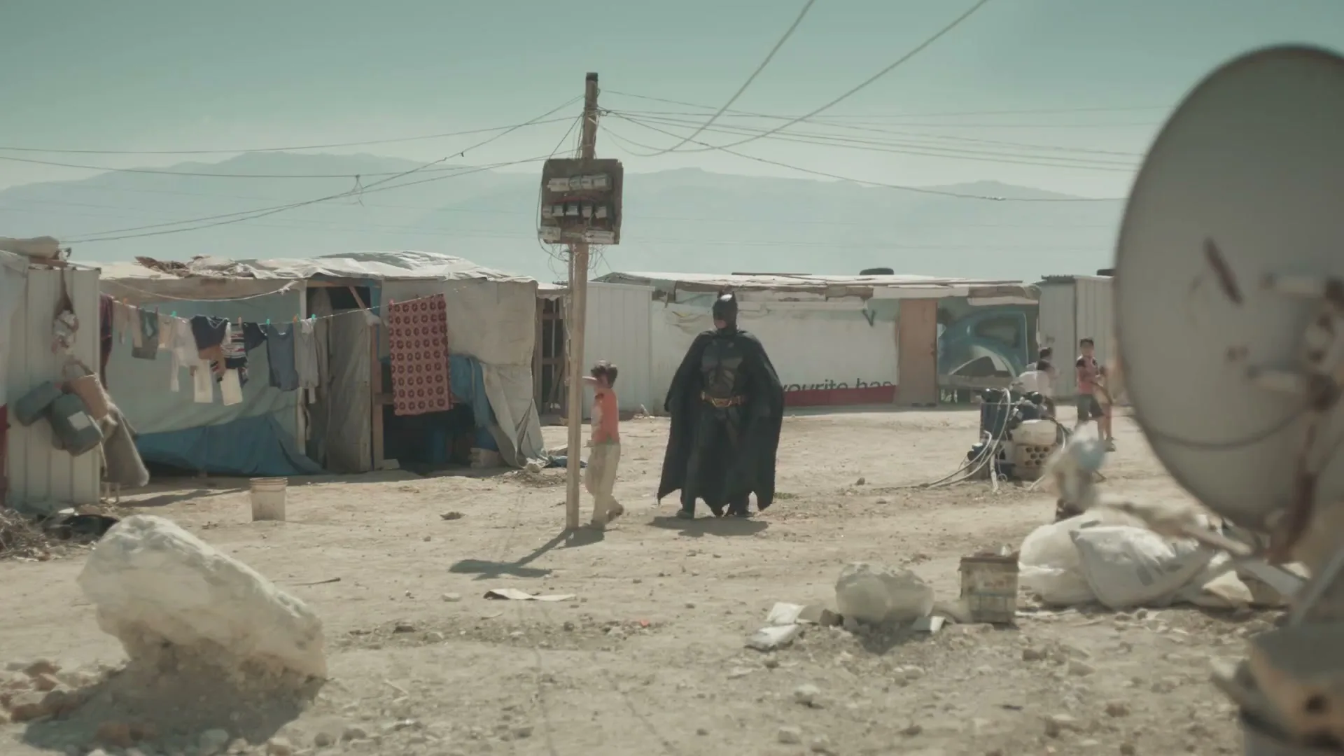 A child in a light orange shirt and light pants walks next to a person dressed as Batman in a refugee camp under a clear sky.
