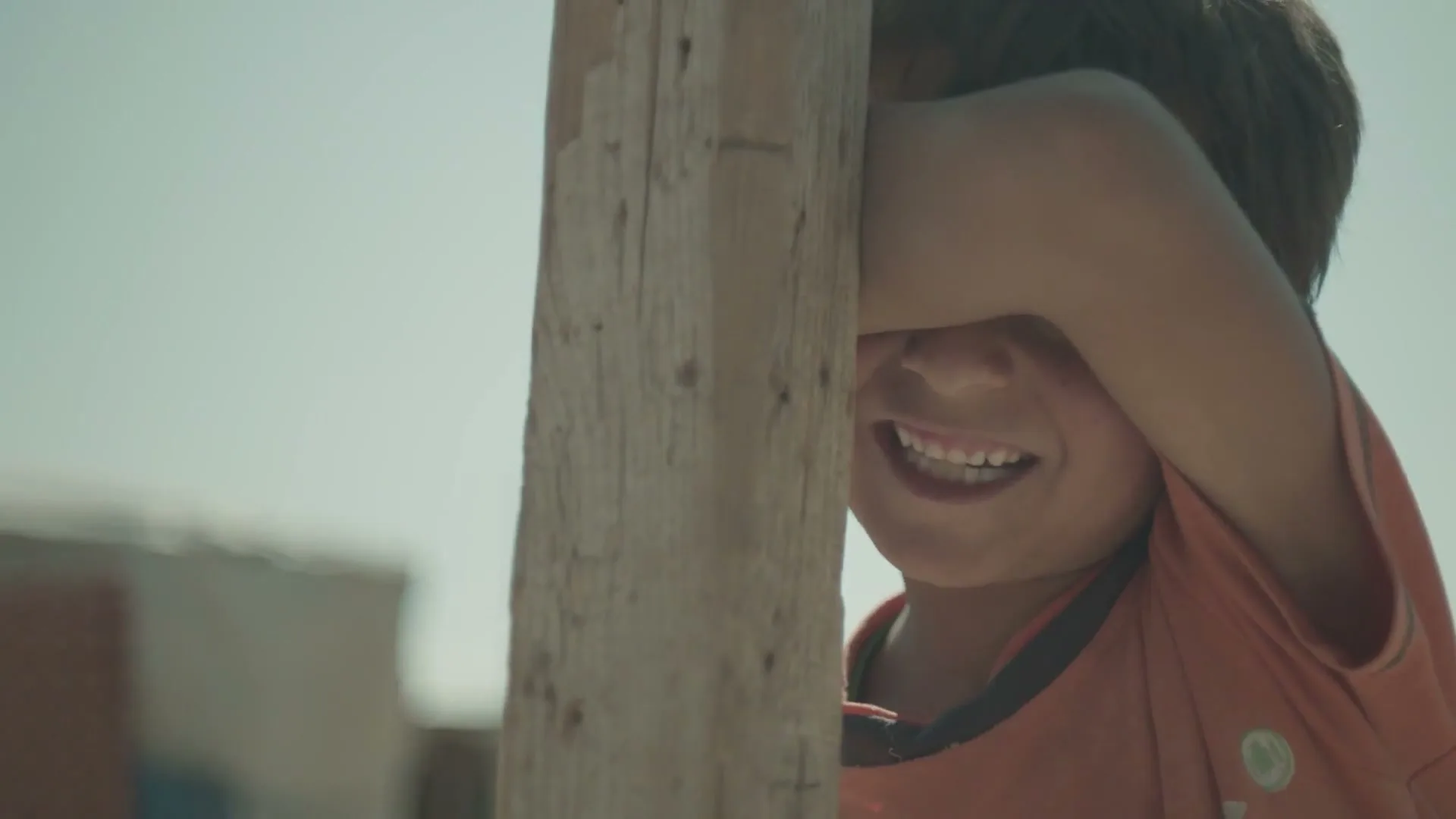 A young boy with dark hair and an orange shirt peeks from behind a wooden post, smiling playfully in an outdoor setting.