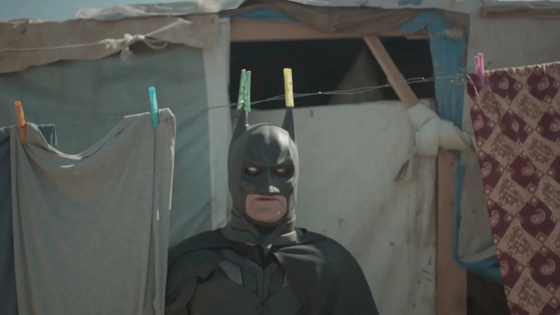 A man dressed as Batman stands in front of a makeshift tent with laundry hanging on a line, looking around with a serious expression.