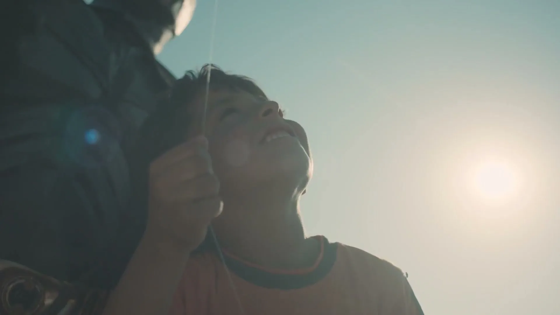 A young boy, wearing an orange t-shirt, looks up with a happy expression, holding a kite string against a bright, sunny sky.