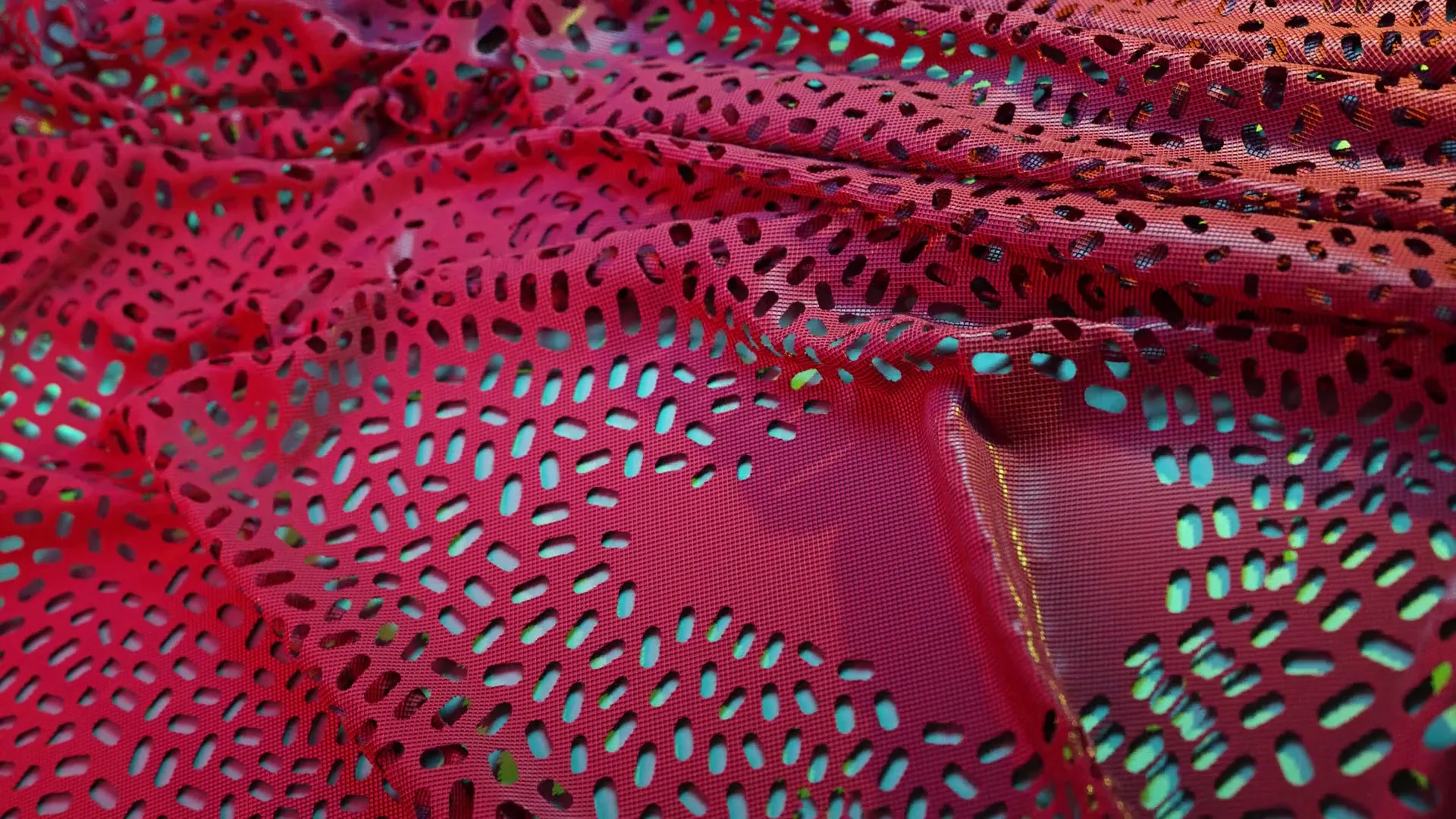 A close-up shot of a vibrant red fabric with small, irregular holes, revealing a shimmering turquoise layer underneath, creating a dynamic visual texture.