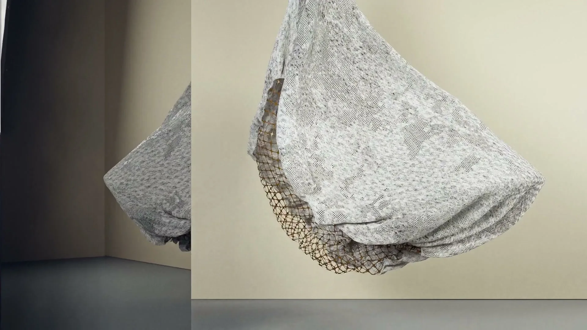 A textured, grey, abstract object with a brown net-like underside floats and undulates in a minimalist room with a reflection.