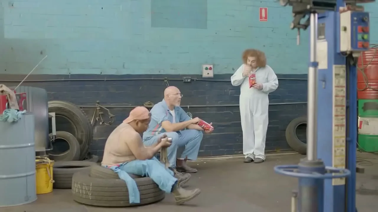 Three mechanics are on break in a garage, two sitting on tires and one standing, all eating Doritos chips.