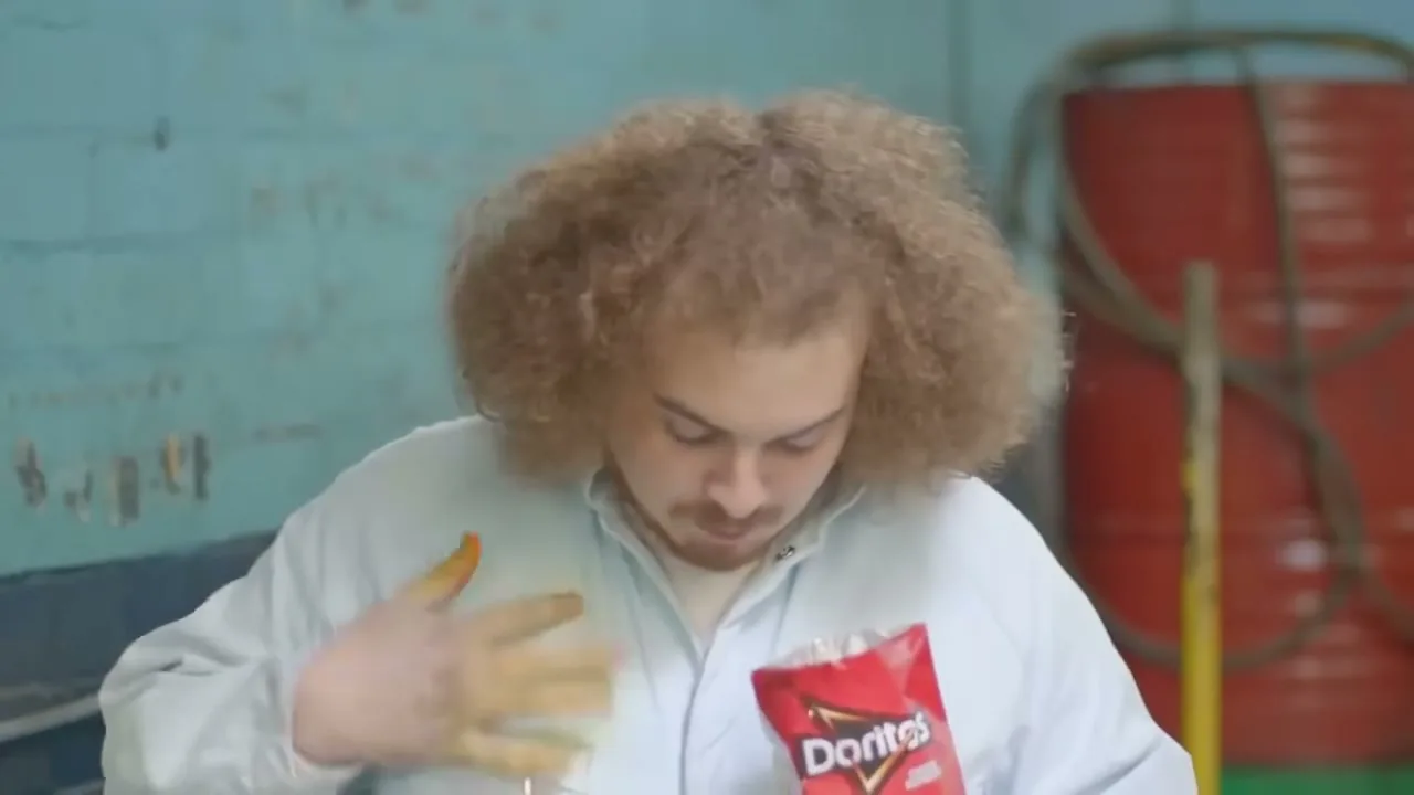A young man with a large afro eats Doritos, then looks at his cheesy fingers, in a garage.